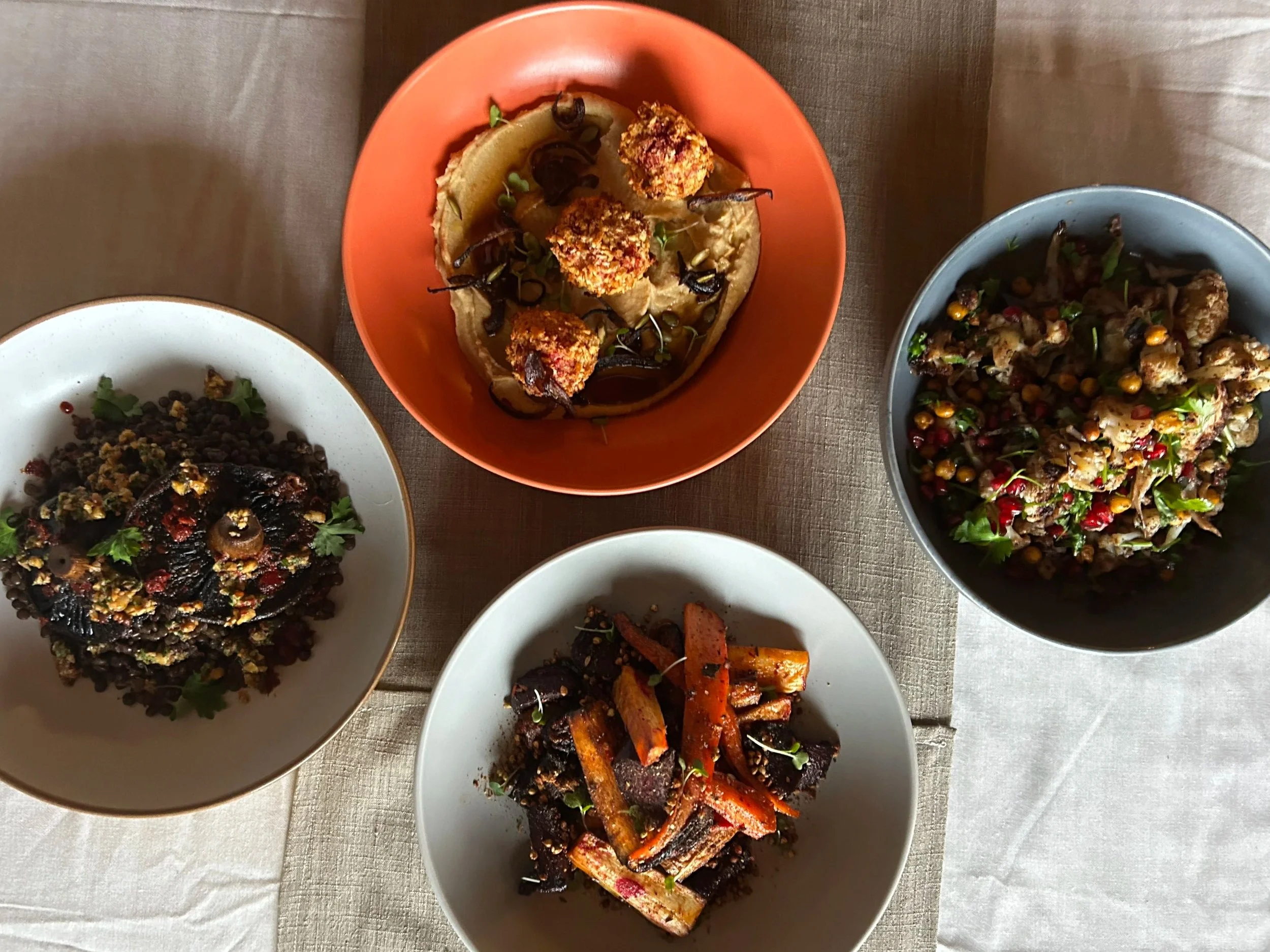 Top-down view of five bowls of diverse colorful dishes on a beige tablecloth.