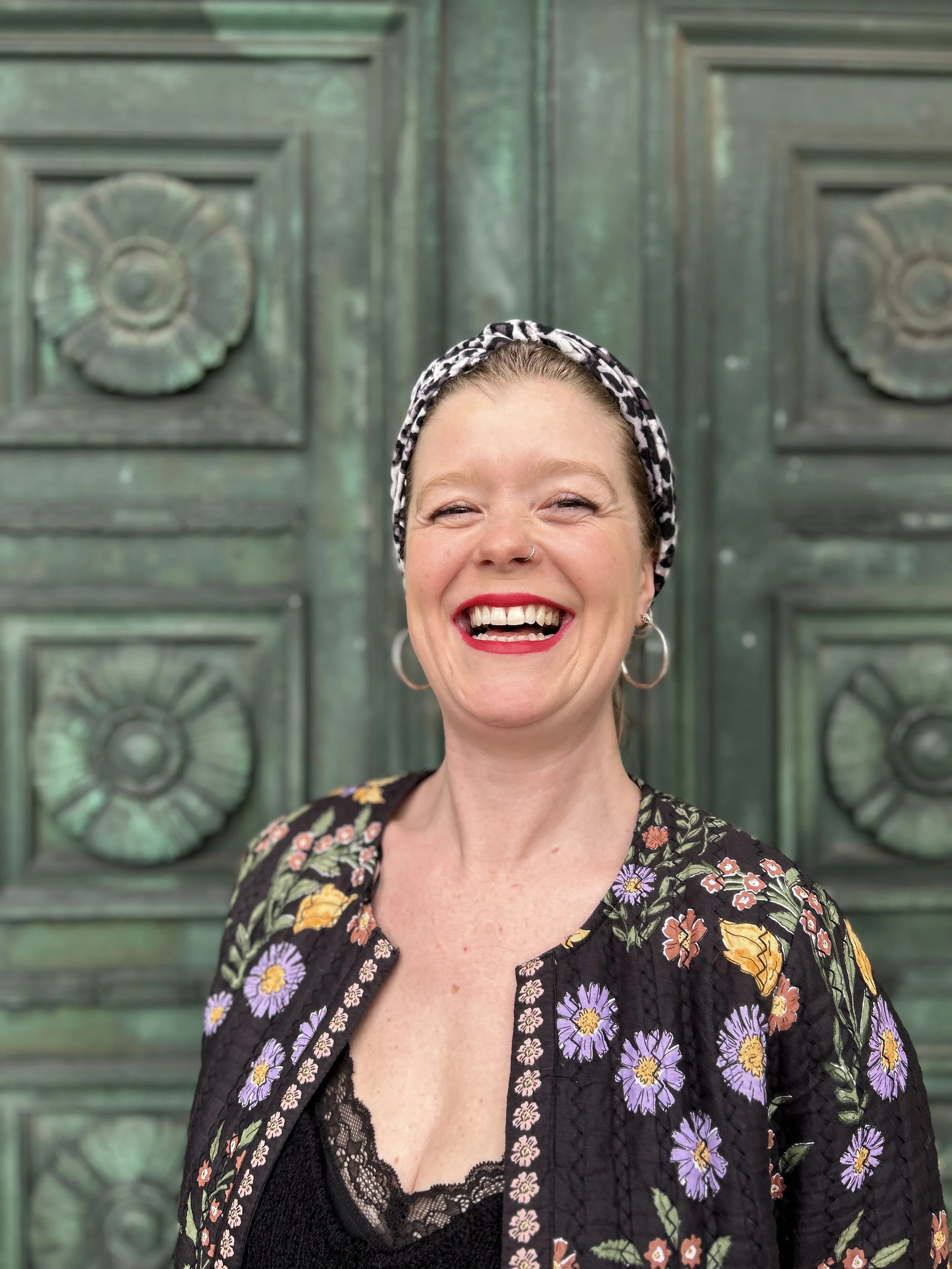 A woman smiling broadly in front of a textured green metal background with circular decorative patterns.