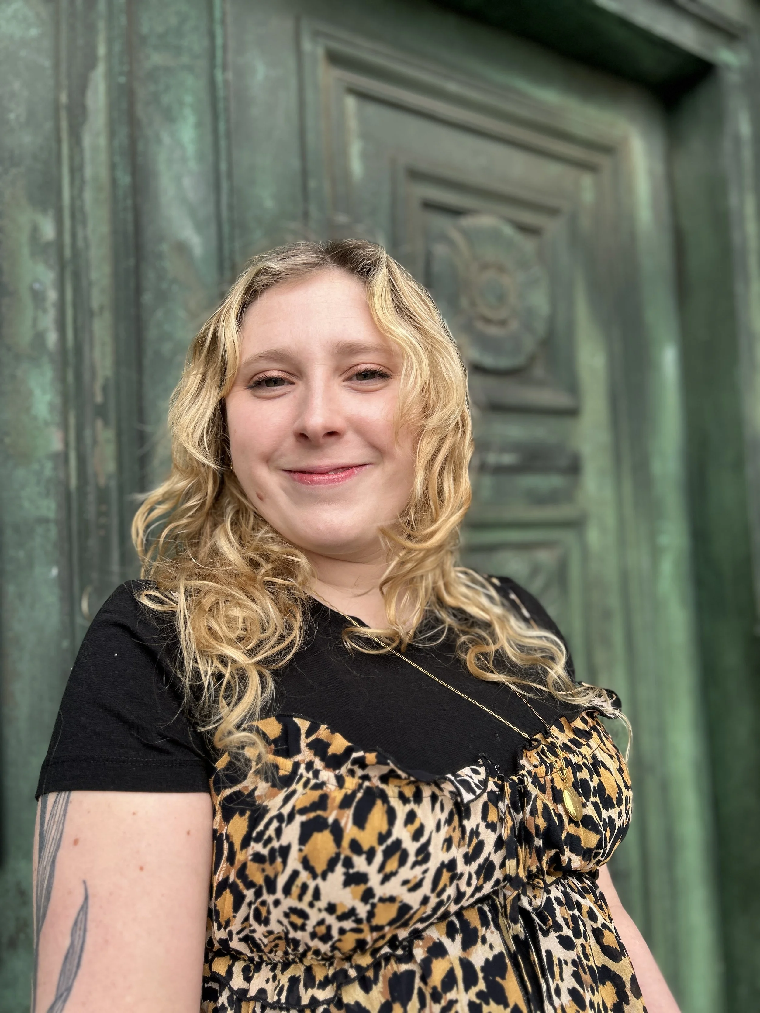 A woman with curly blonde hair smiling at the camera, wearing a black and leopard print dress with a gold necklace, standing in front of a vintage green door.
