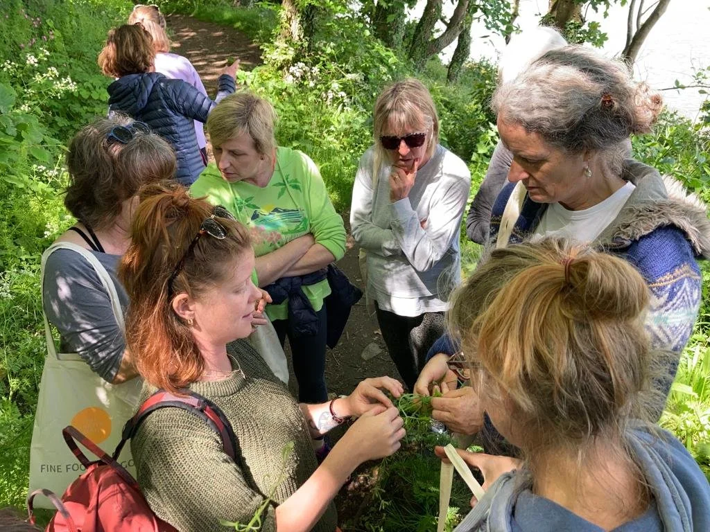 A group of women gathered outdoors on a trail surrounded by greenery, examining and discussing plants.