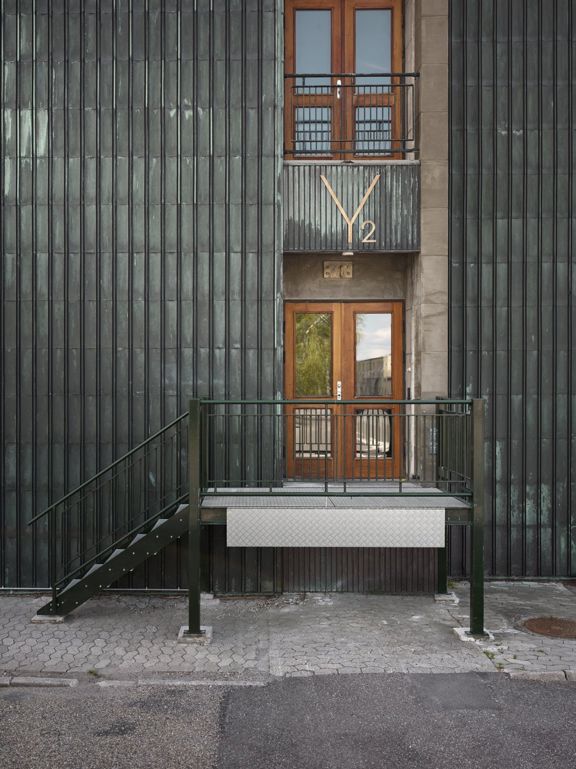 A modern building facade with dark green vertical metal panels, wooden framed glass doors, and balconies with black metal railings. A large wall clock displays 'Y2' in gold.