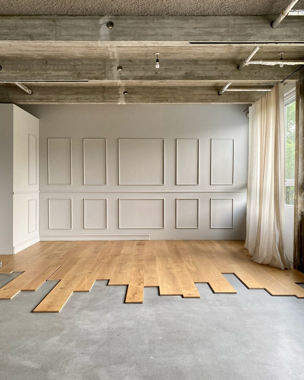 Interior of a room under renovation with a partially installed hardwood floor, ceiling with exposed pipes and concrete, a white wall with decorative molding, a window with sheer curtains, and an unfinished floor area.