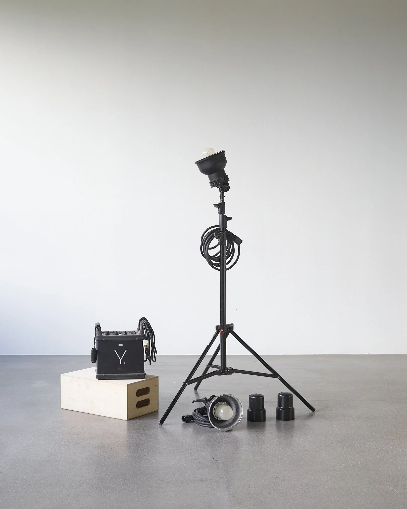 Studio photography equipment including a large tripod with light, a light stand, a bag of cables, a spotlight, and some light modifiers on the floor against a plain wall.