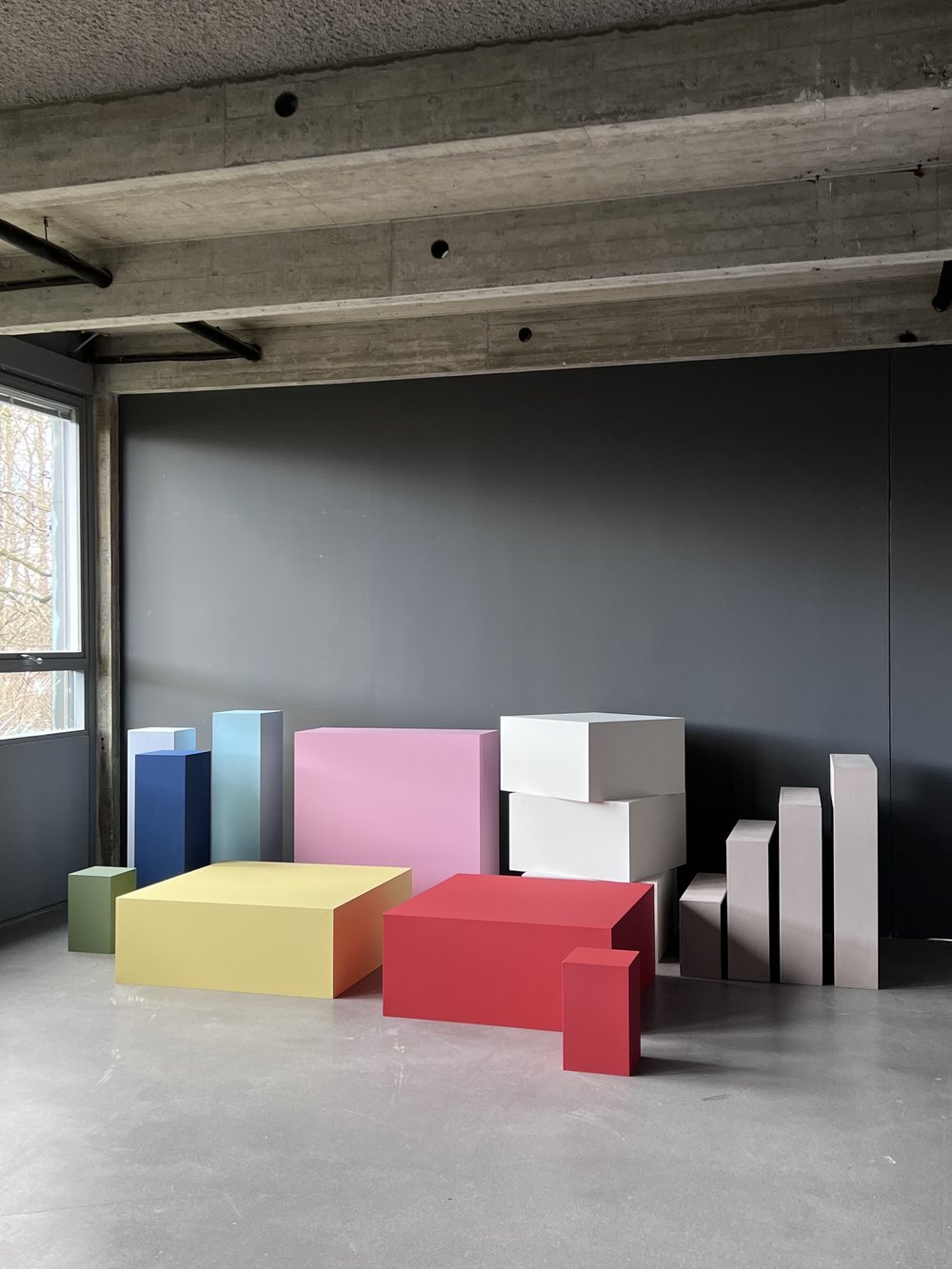 Colorful geometric blocks resembling a bar chart arrangement in a modern industrial-style room with concrete ceiling and dark wall, next to a window.