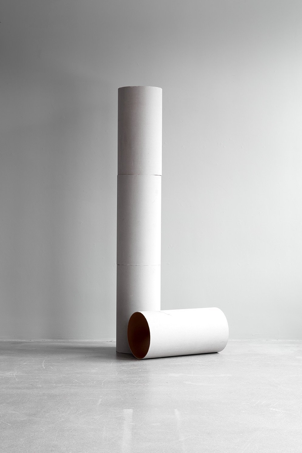 Three large white cylindrical tubes, two standing vertically and one lying horizontally, against a plain light gray wall and floor.