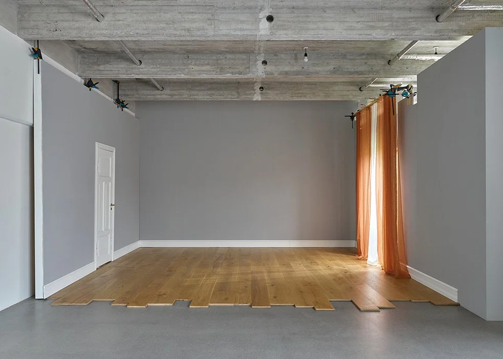 Empty room with gray walls, exposed concrete ceiling, partially installed hardwood flooring, and orange curtains covering a window.