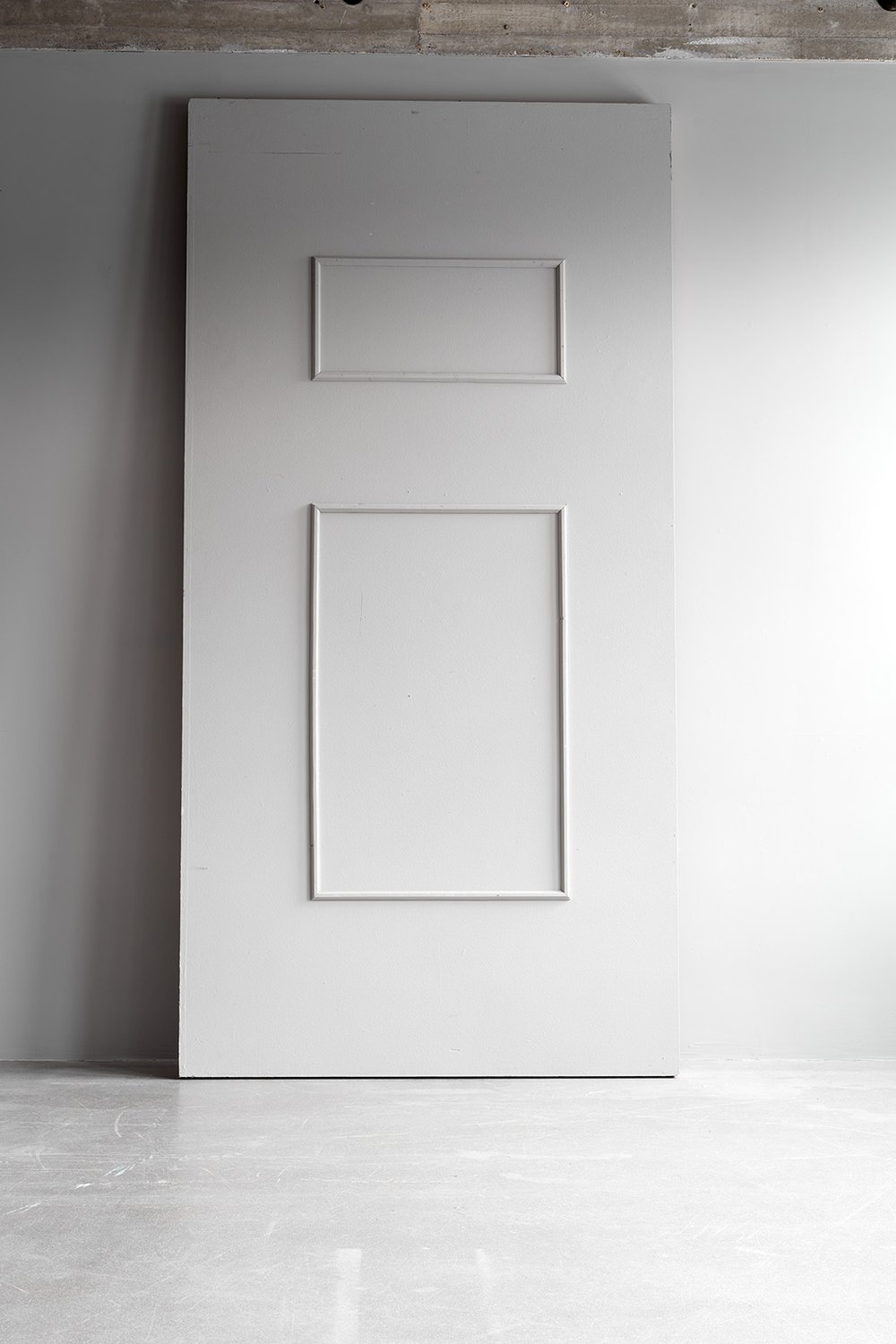 White door with two rectangular panels, leaning against a wall in a room with light flooring and partly visible ceiling.