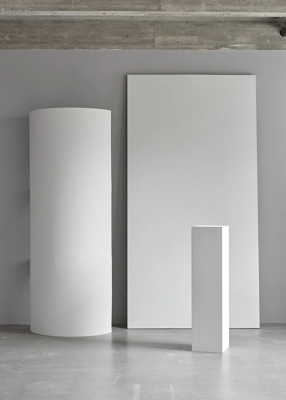 Minimalist art installation with a white cylindrical sculpture, a tall rectangular white panel, and a smaller white rectangular pedestal against a gray wall and concrete floor.