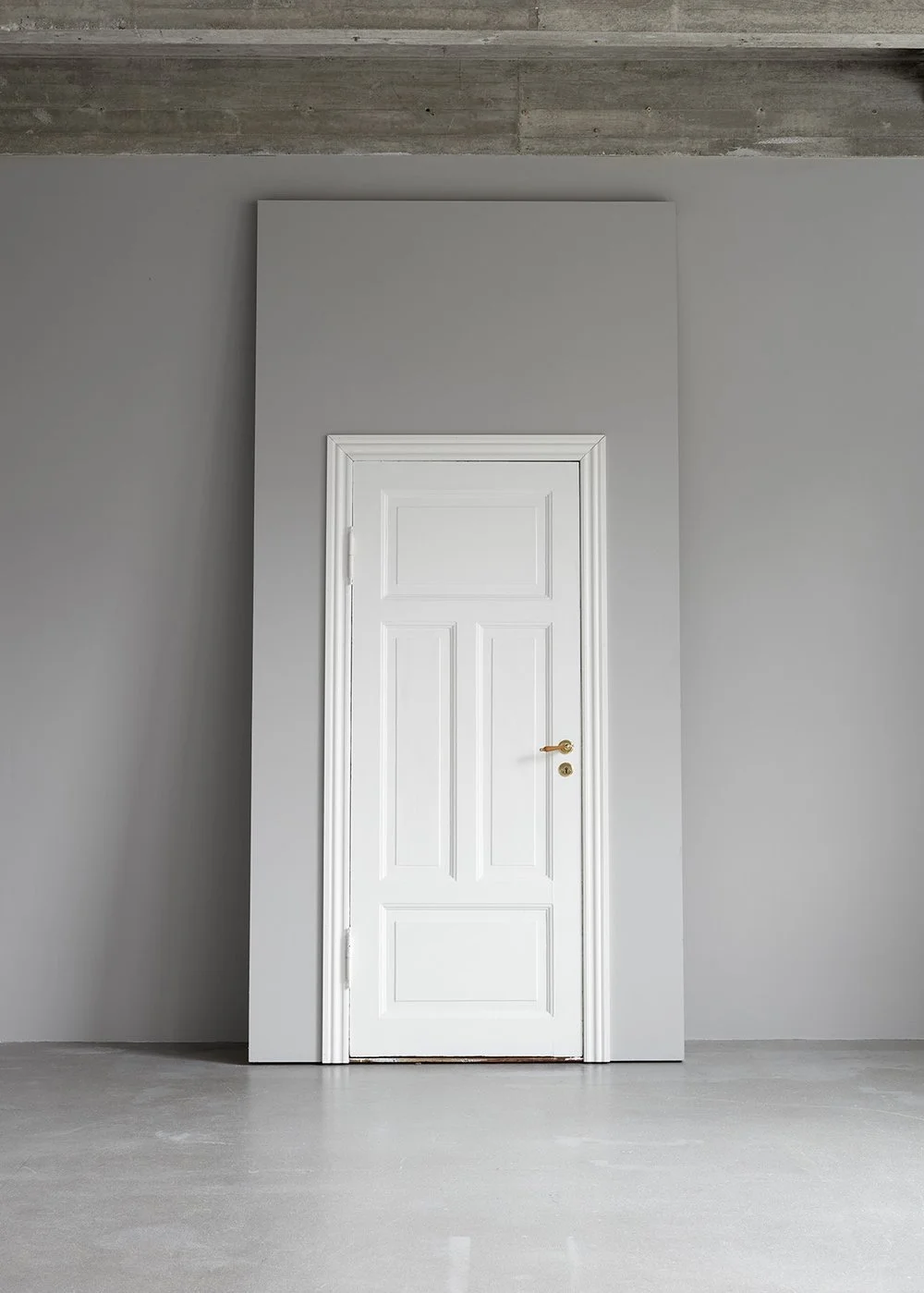 White door with brass handle and lock set, framed by white door casing, set in a plain gray wall in an unfinished room with concrete floor and exposed concrete ceiling.