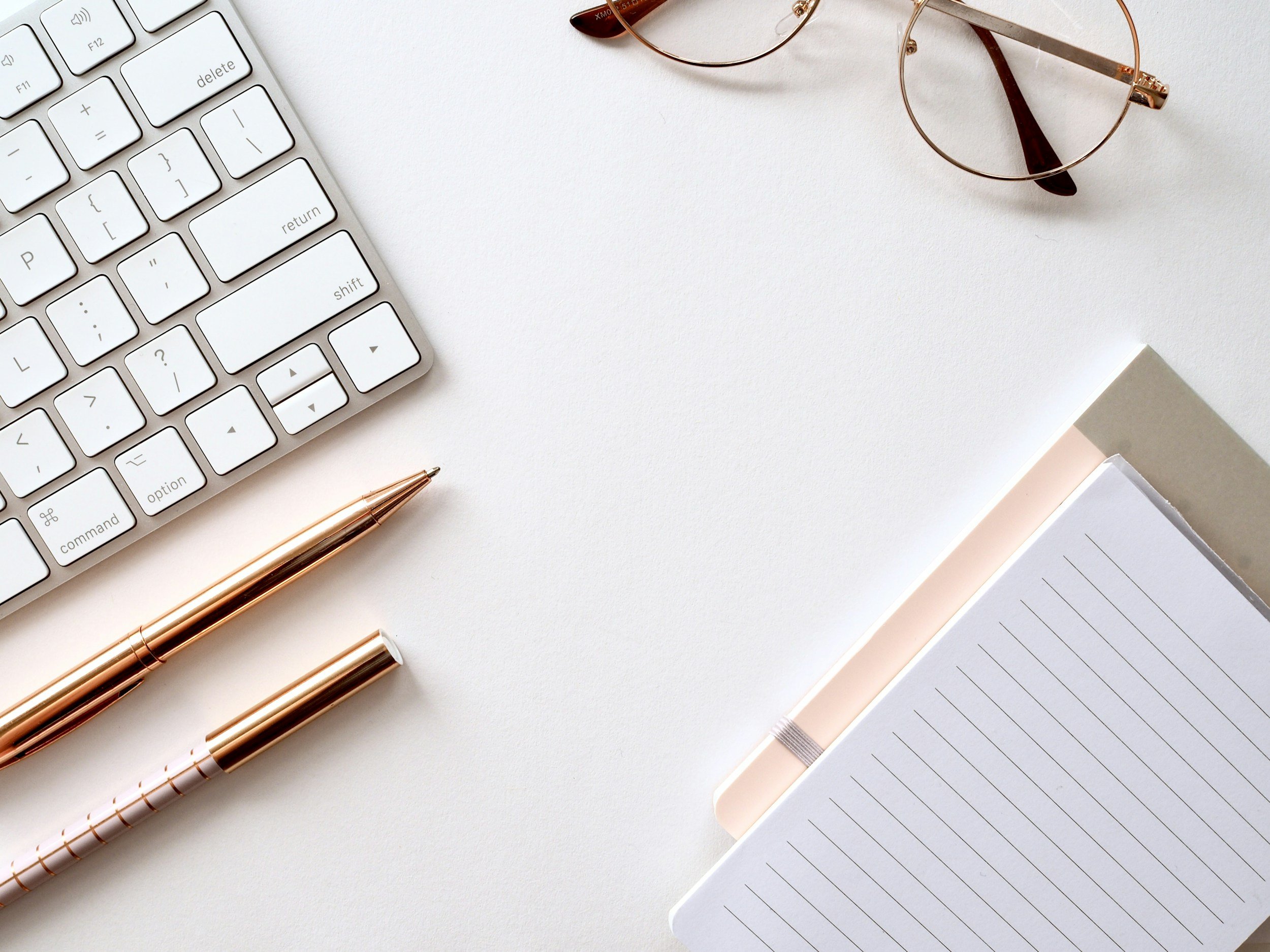 White desk with a partial white keyboard, two rose gold pens, a pair of round eyeglasses, and a lined notebook.