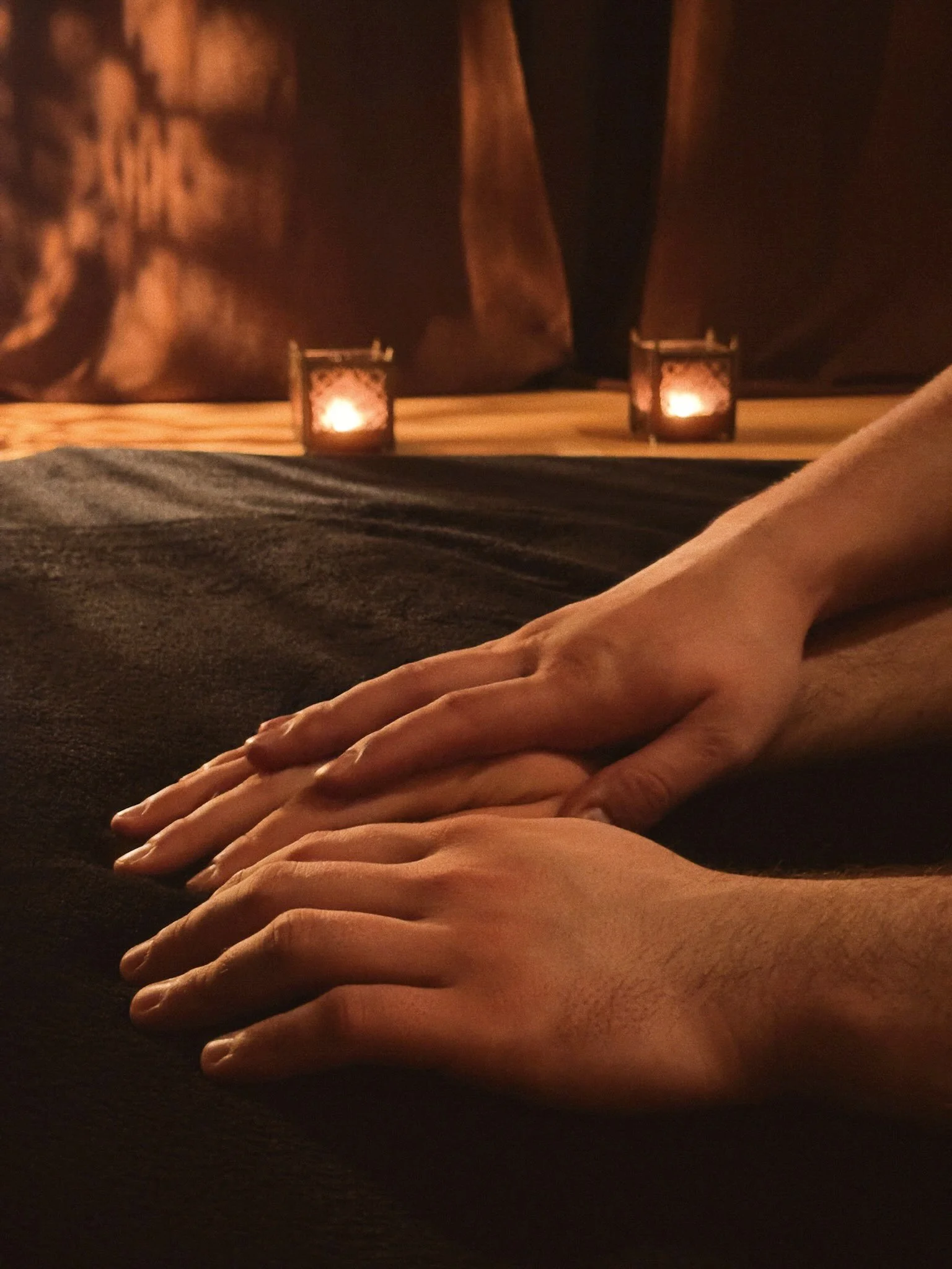 Close-up of two people's hands resting on a dark surface, with a warm, dimly lit setting and two small lanterns in the background.
