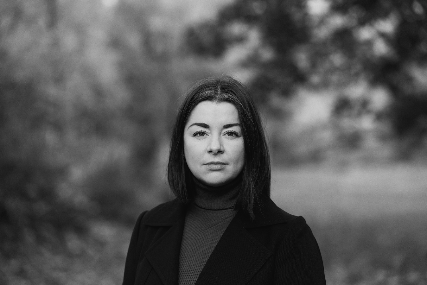 Black and white portrait of Suzanne Rietmeijer, a woman, with shoulder-length dark hair, wearing a turtleneck and a blazer, standing outdoors with trees and blurred nature in the background.