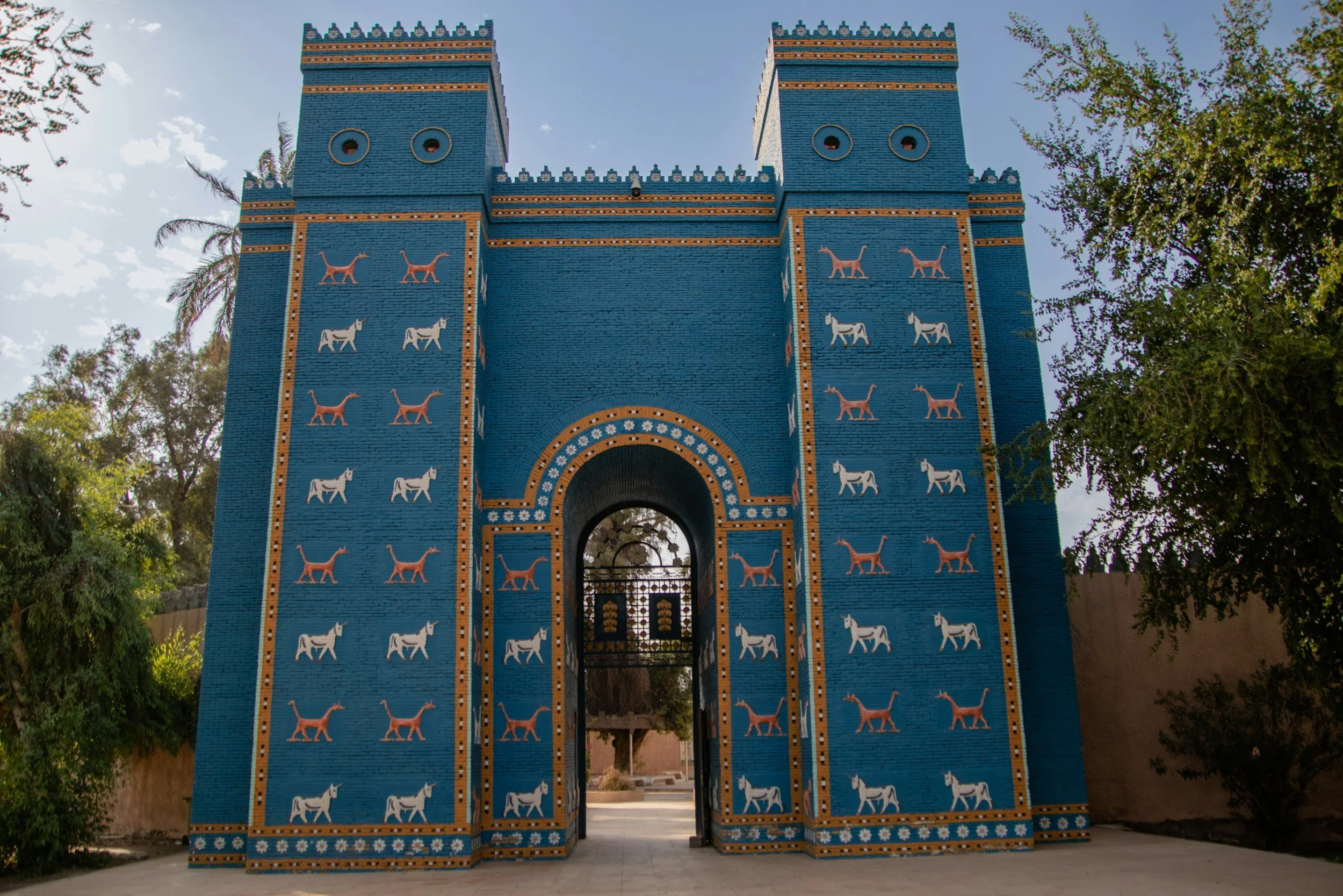 A large, colorful brick gateway painted bright blue with orange and white decorative patterns, featuring illustrations of donkeys and camels, and topped with crenellations. The gate has an arched opening at the bottom and is surrounded by trees and a partly cloudy sky.