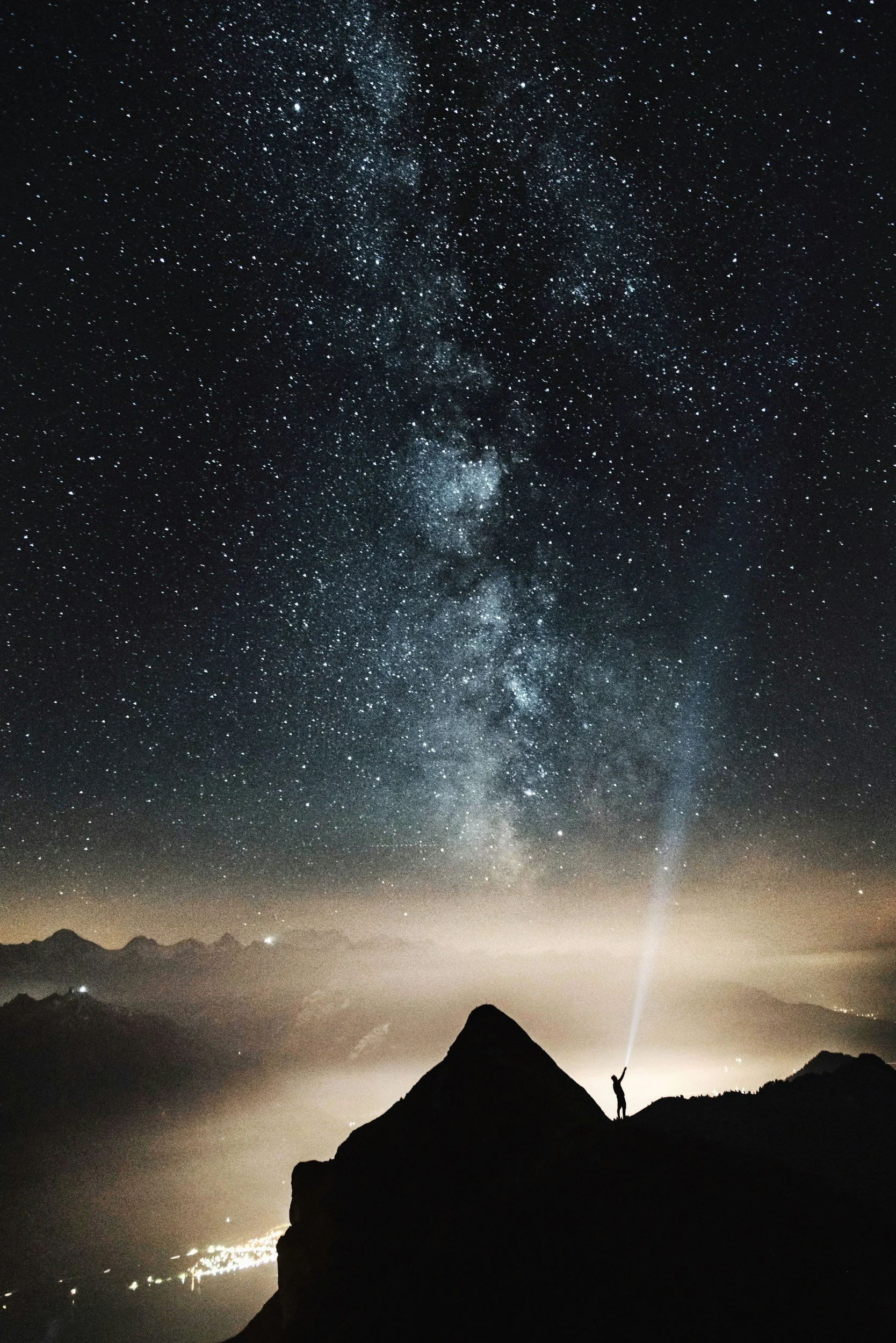 A person stands on a mountain peak at night, looking up at the star-filled sky and the Milky Way galaxy, with a flashlight beam pointing downwards.