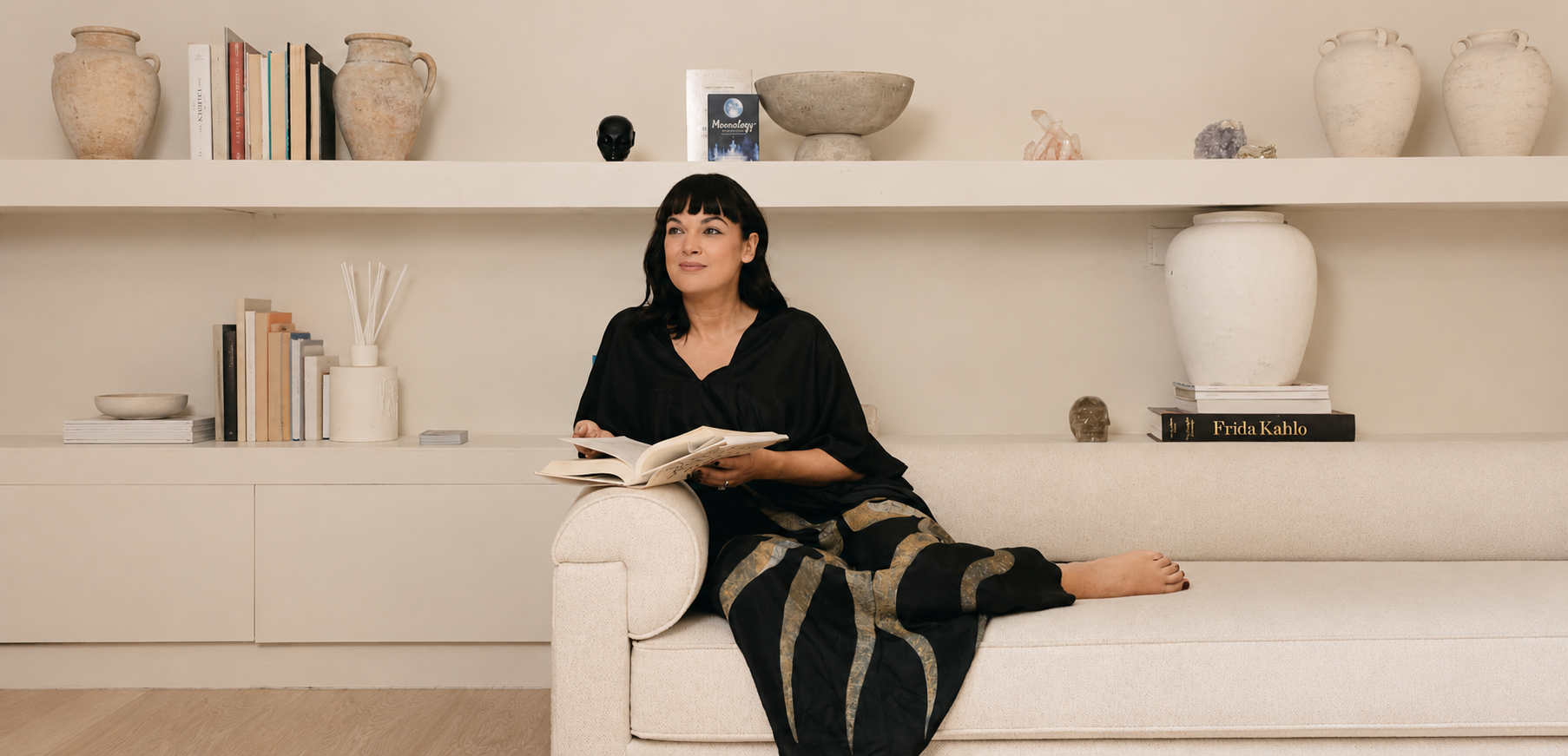 Rachael Hall photographed at home, seated on a cream sofa with an open book amid shelves of books, ceramics and crystals.