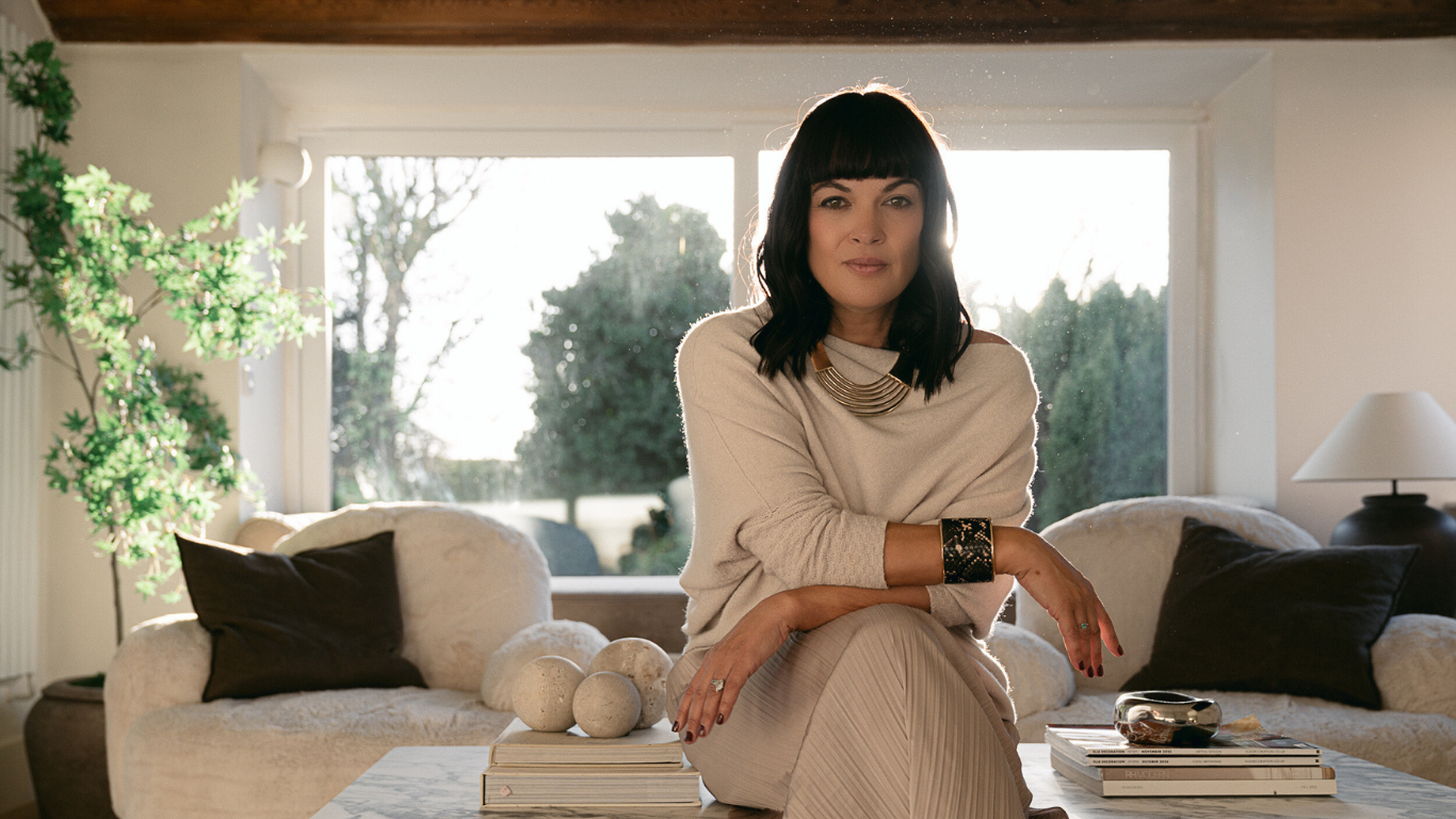 A woman with dark hair and bangs sitting on a coffee table in a living room, with sunlight streaming through a large window behind her. The room has cream-colored sofas with dark pillows, a lamp, a green plant, and decorative objects on the table.