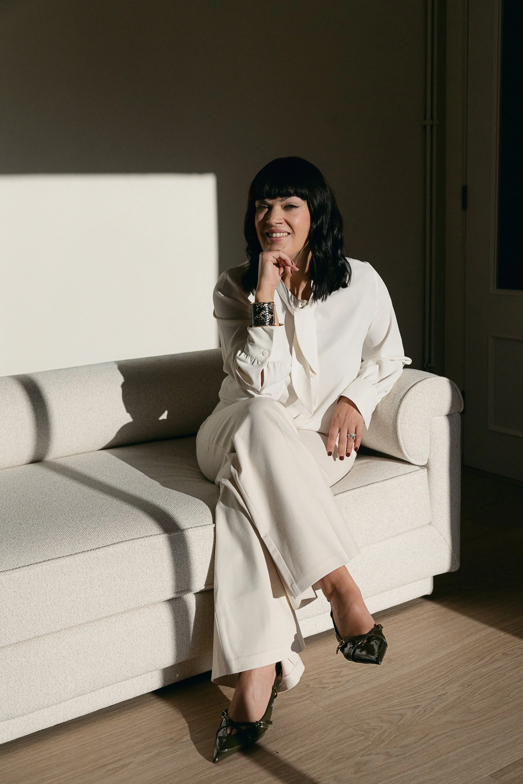 A woman with black hair, wearing a white suit, sitting on a white couch, smiling, with one leg crossed over the other, and wearing black high-heel shoes, in a room with dark floors and walls.