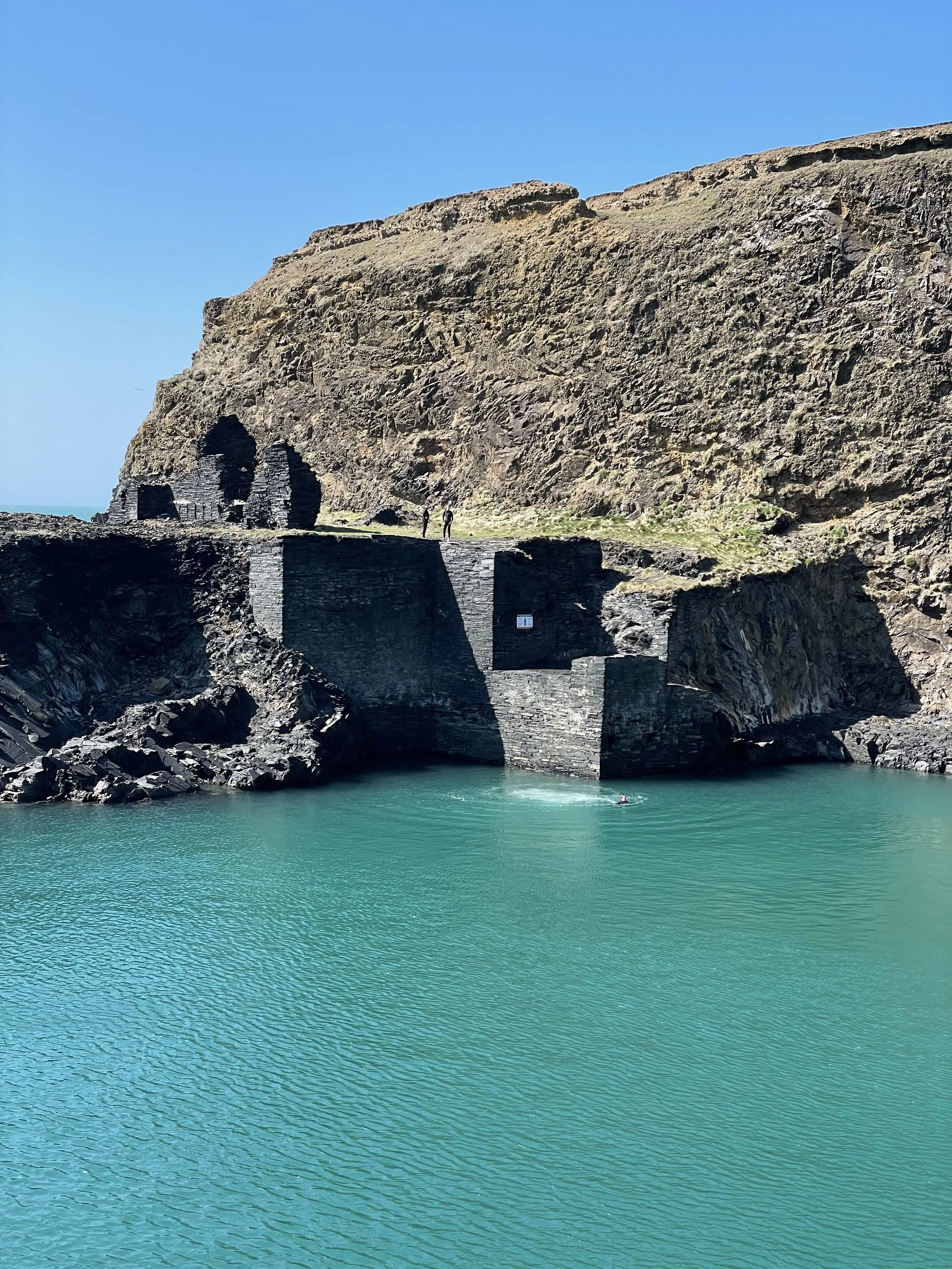 A coastal scene with turquoise water, rugged rocky cliffs, and an old stone structure built into the cliffside under a clear blue sky.