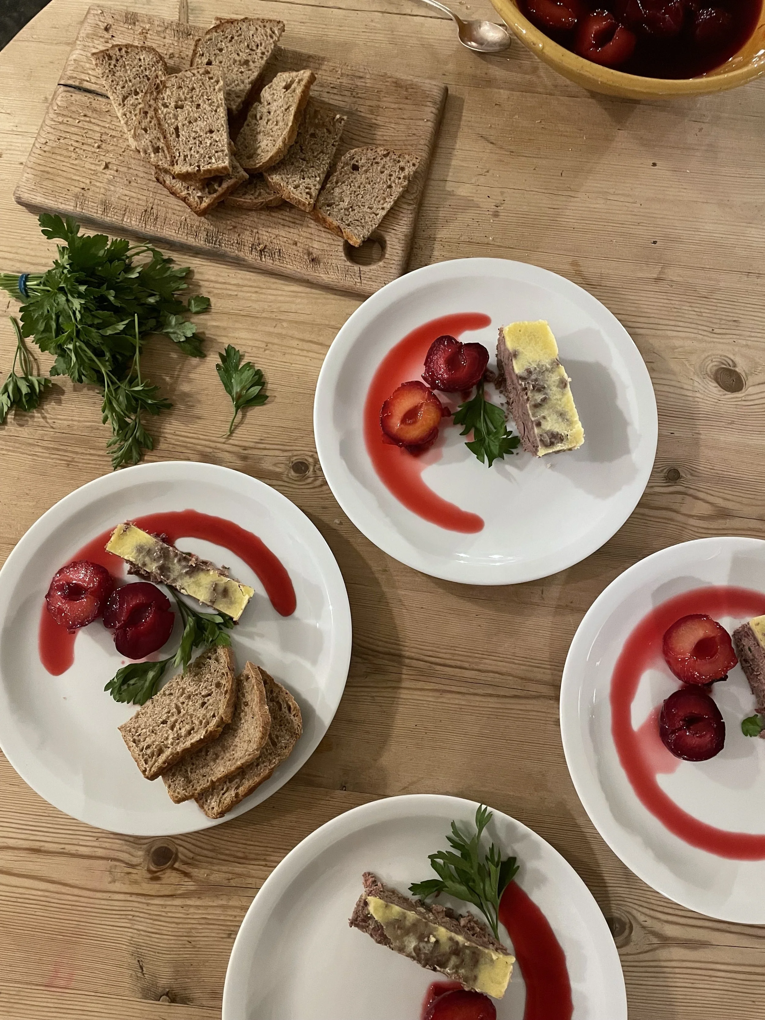 Three small white plates with dessert slices, strawberry sauce, and fruit garnishes, placed on a wooden table. A wooden board with bread slices, a bowl of cherries, and sprigs of parsley are also visible.