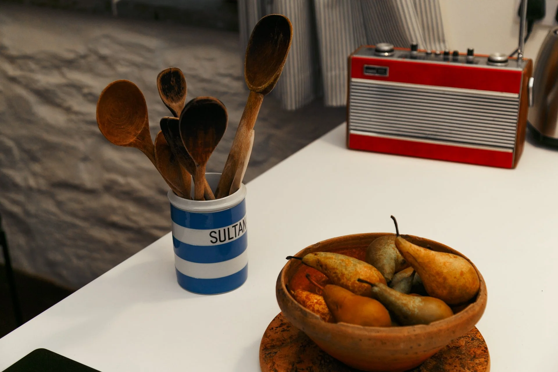 Wooden spoons in a blue and white striped container labeled 'SULTAN' on a white surface, with a bowl of pears and a retro red and gray radio in the background.