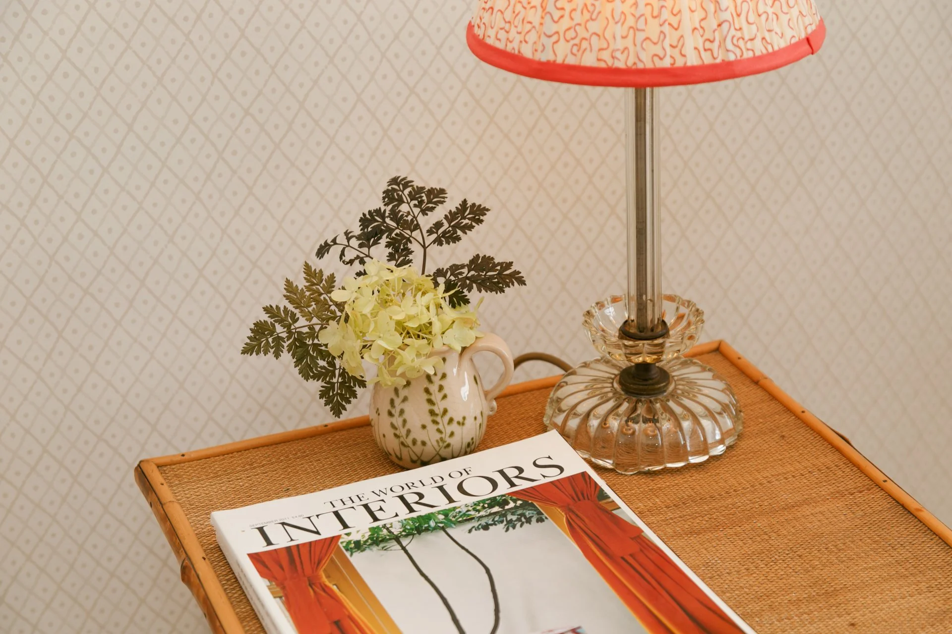 A wooden table with a magazine titled 'The World of Interiors,' a ceramic mug with green leaf design and white flowers, and a glass lamp with a red and white patterned lampshade against a patterned wall.