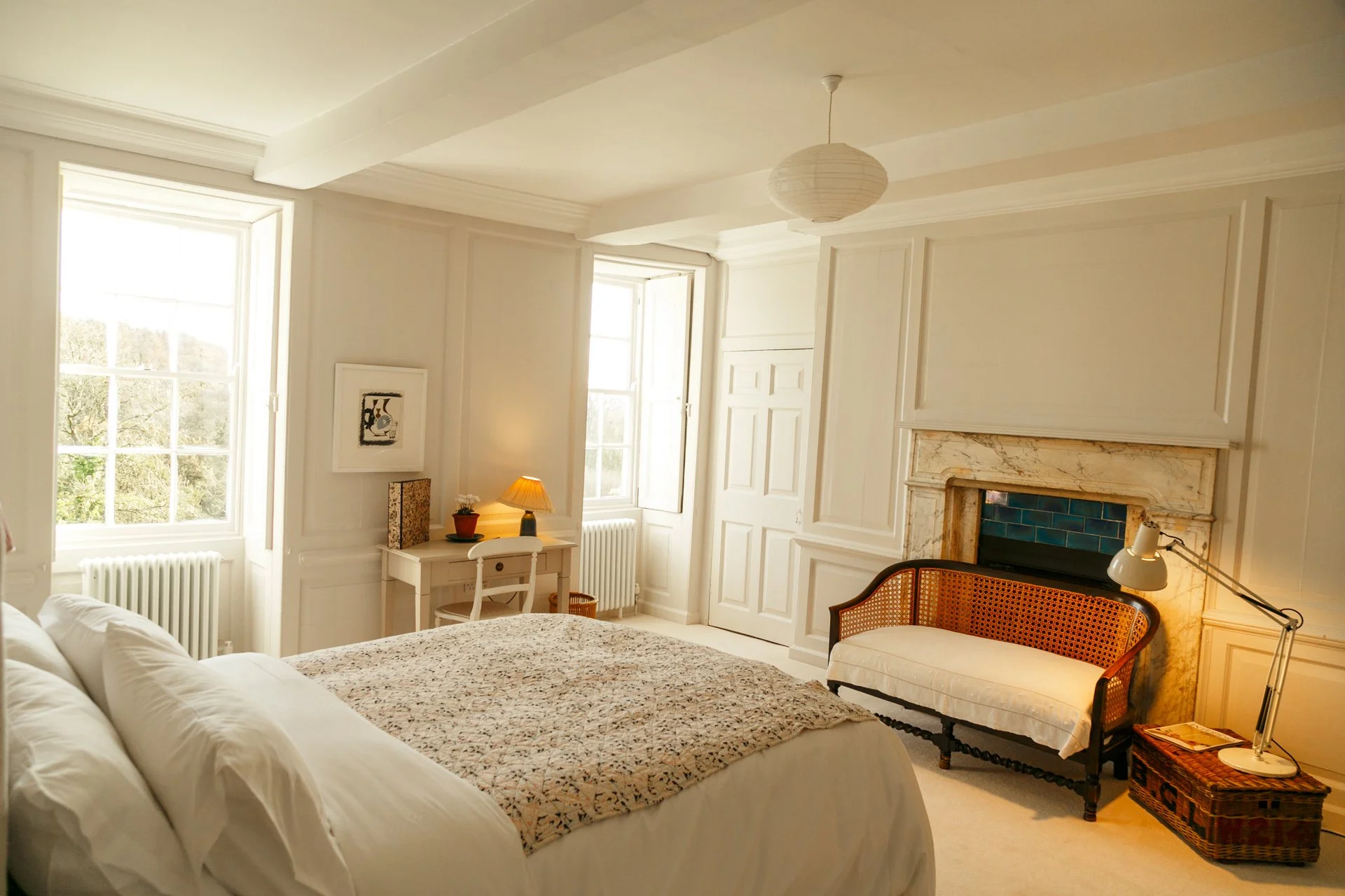 Bright bedroom with large windows, a white bed with a patterned bedspread, a vintage side chair, a fireplace with marble surround, a small desk with a lamp, and a wicker trunk.