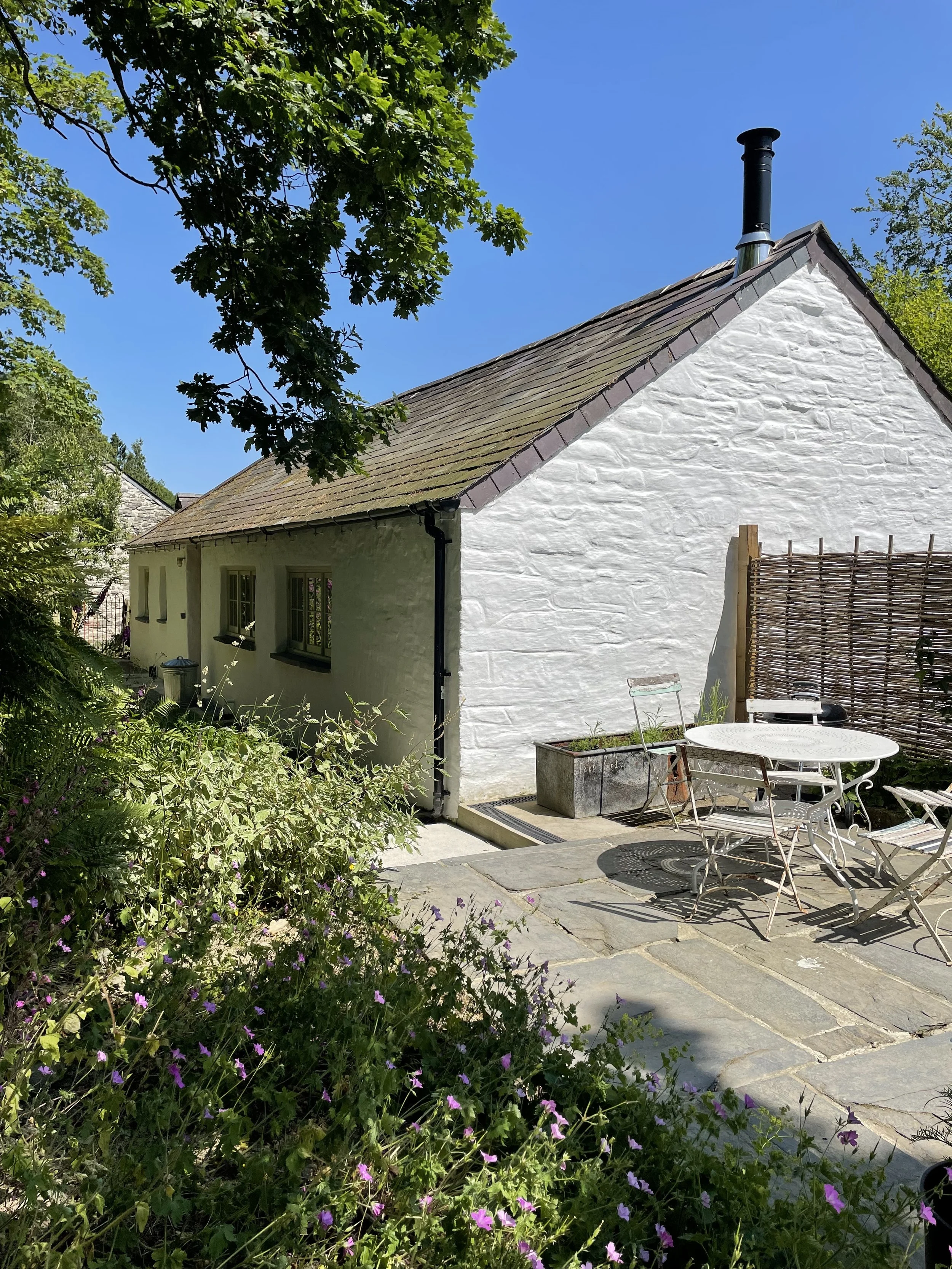 A white cottage with a tiled roof and chimney, surrounded by greenery and flowers, with a patio area featuring a round table and chairs.