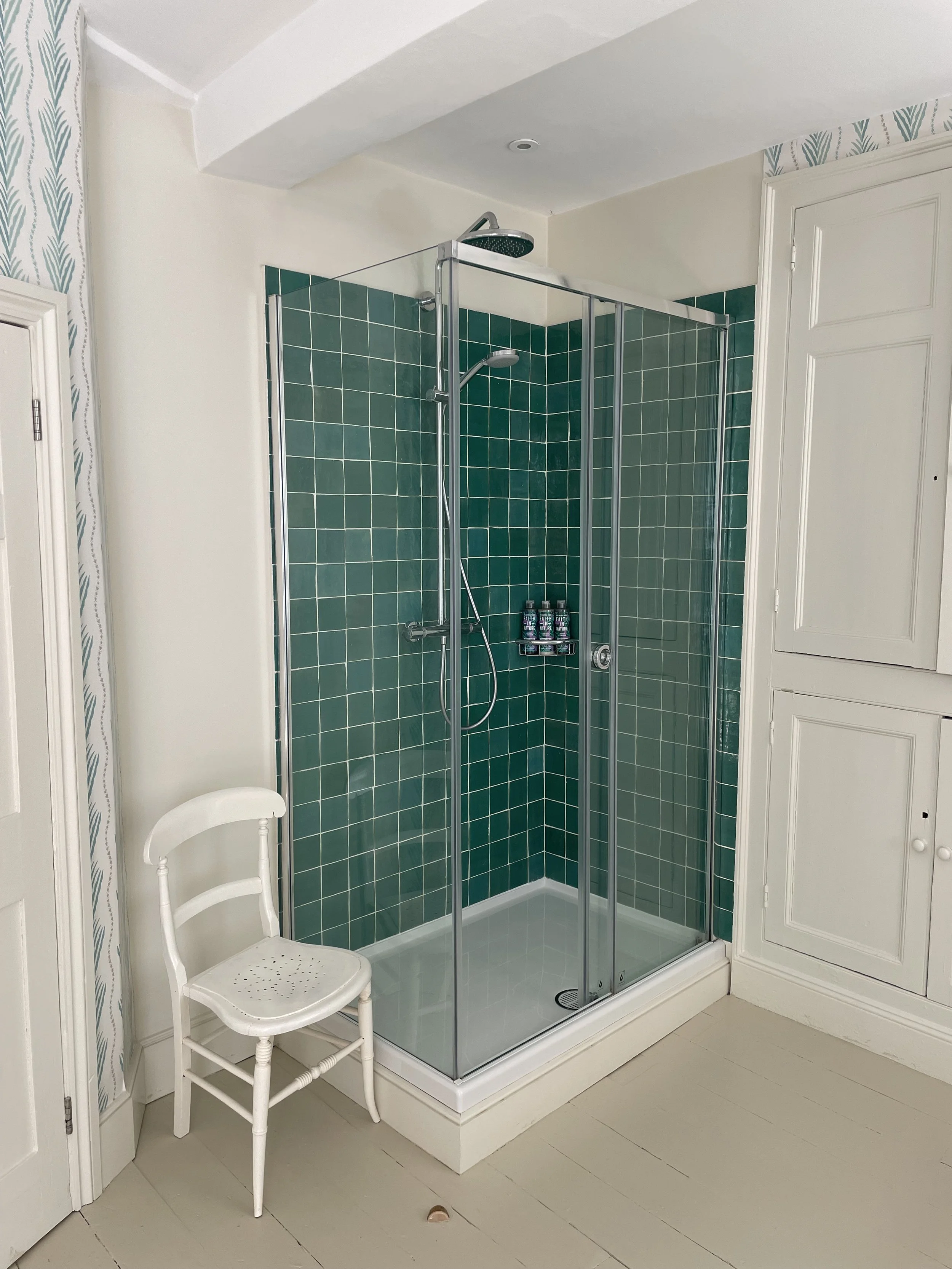 A small bathroom shower with green tiled walls, a glass door, a rain shower head, and a handheld shower, with a white chair beside it and white cabinetry nearby.