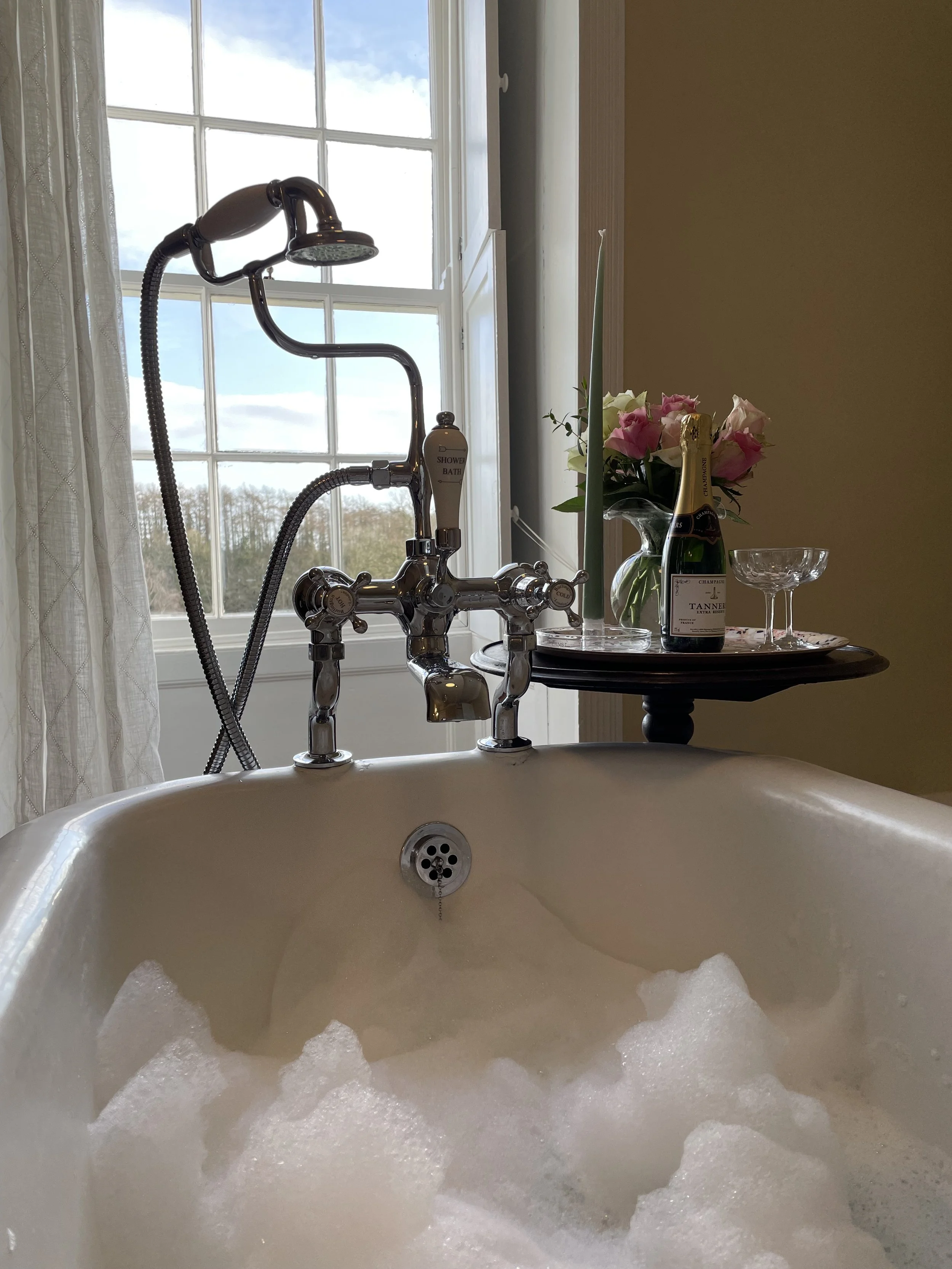 A bathtub filled with bubbles in a room with a large window, a side table with a bouquet of pink and white roses, a bottle of champagne, two glasses, and a single candle.