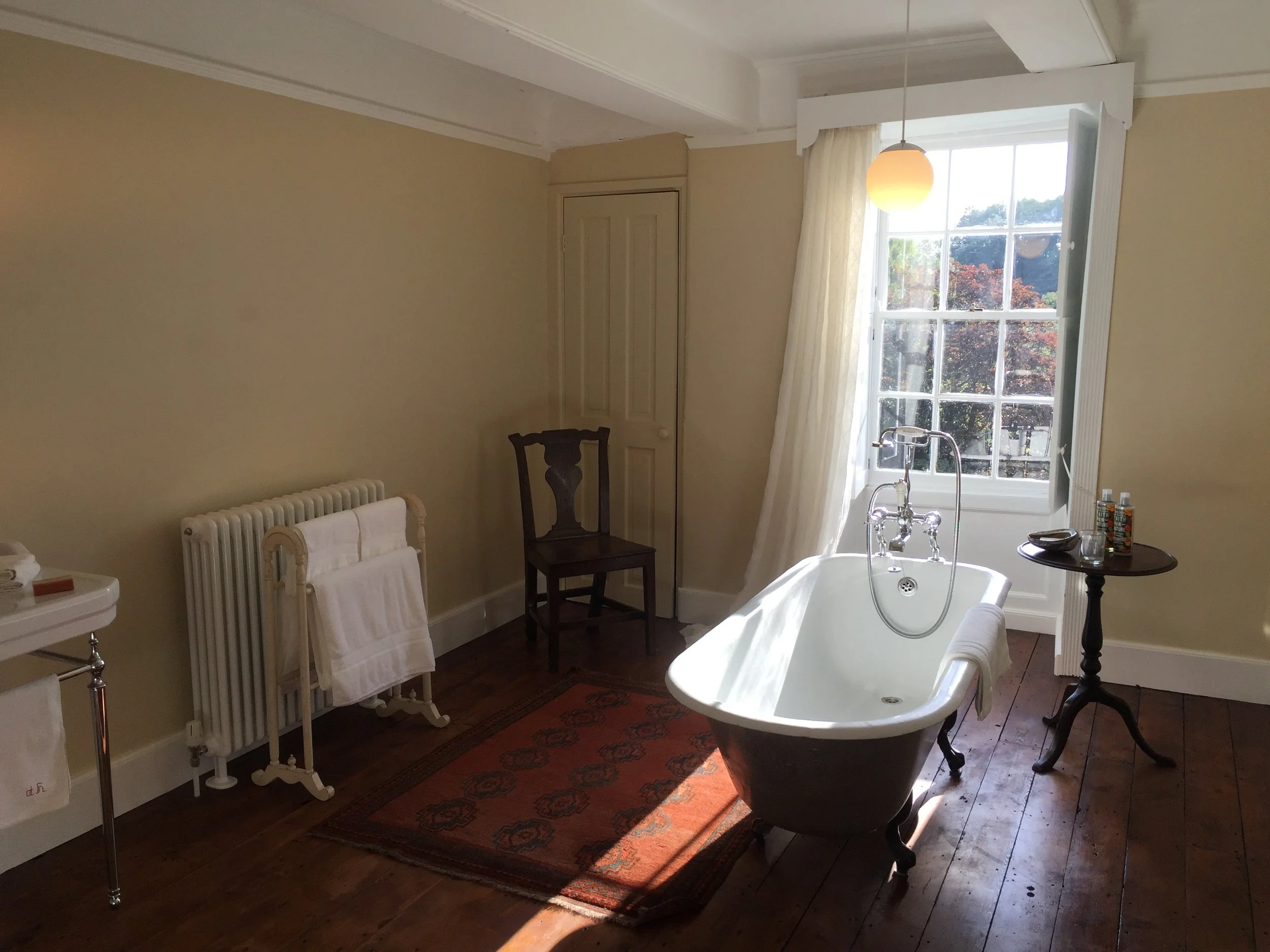 Bathroom with a vintage clawfoot tub, a window with natural light, a small round table, a dark wooden chair, a radiator with towels, and a patterned rug on wooden floor.