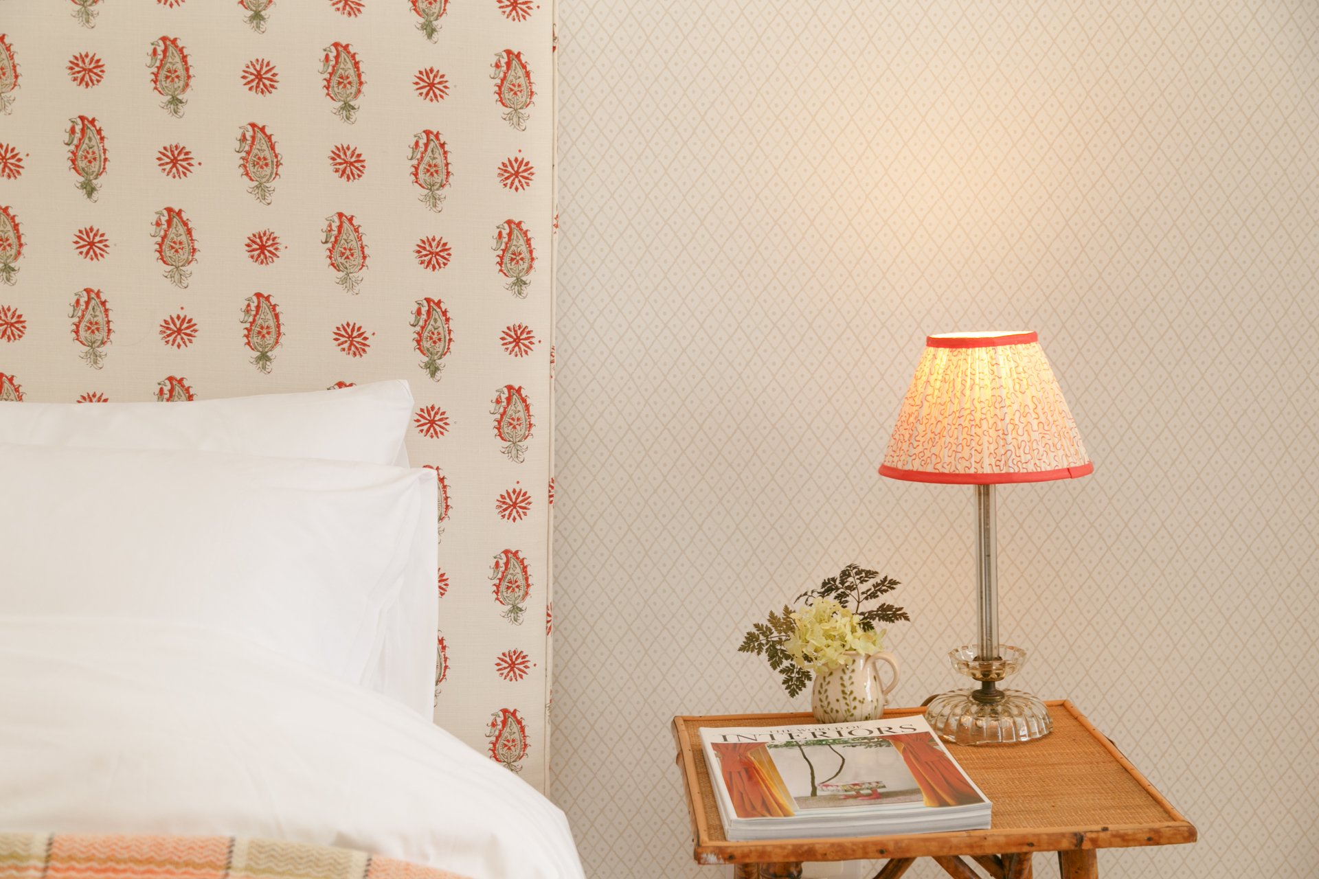 A bedroom with a white bed and pillows, a nightstand with a magazine, a vase with white flowers, and a table lamp, against a wall with patterned wallpaper.