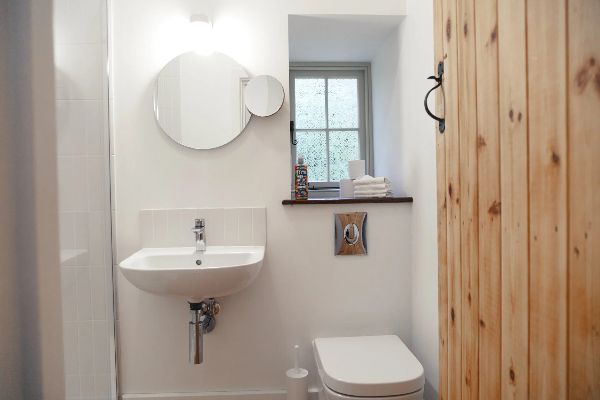 Small bathroom with white walls, a round mirror, a small wall-mounted sink, a window with patterned frosted glass, folded towels, a toilet paper roll, and a toilet, with wood paneling on one wall.