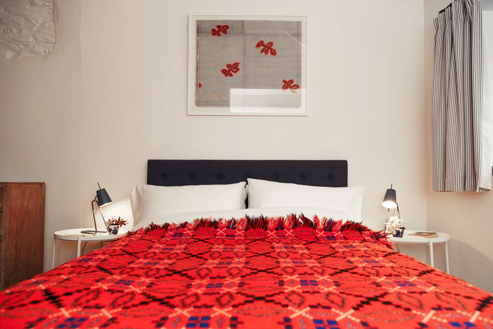 A cozy bedroom featuring a bed with a black headboard, white pillows, and a red patterned blanket. There are two white bedside tables with black lamps, a small plant, and books. A framed artwork depicting red butterflies hangs above the bed, and stri