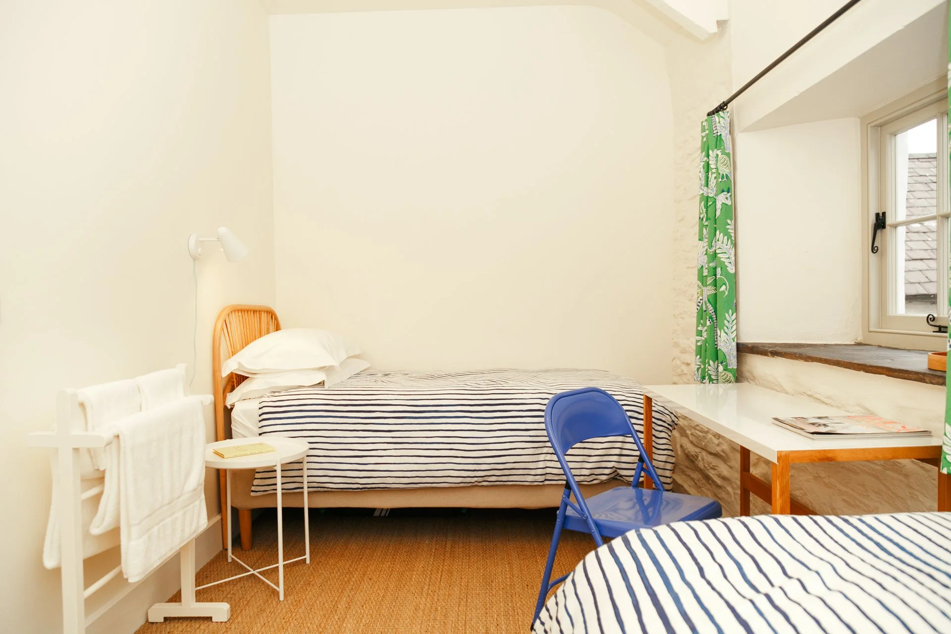 A simple bedroom with two twin beds covered in striped bedding, a blue chair, a small white side table with a book, a window with green leafy patterned curtains, and a white wall with a wall-mounted reading lamp.