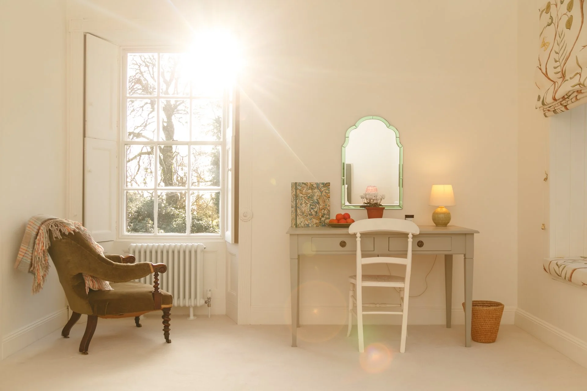 Bright, airy room with a large window letting in sunlight, a vintage armchair with a throw blanket, a small desk with a mirror, a table lamp, fruit bowl, and decorative items, and a woven basket on the floor.