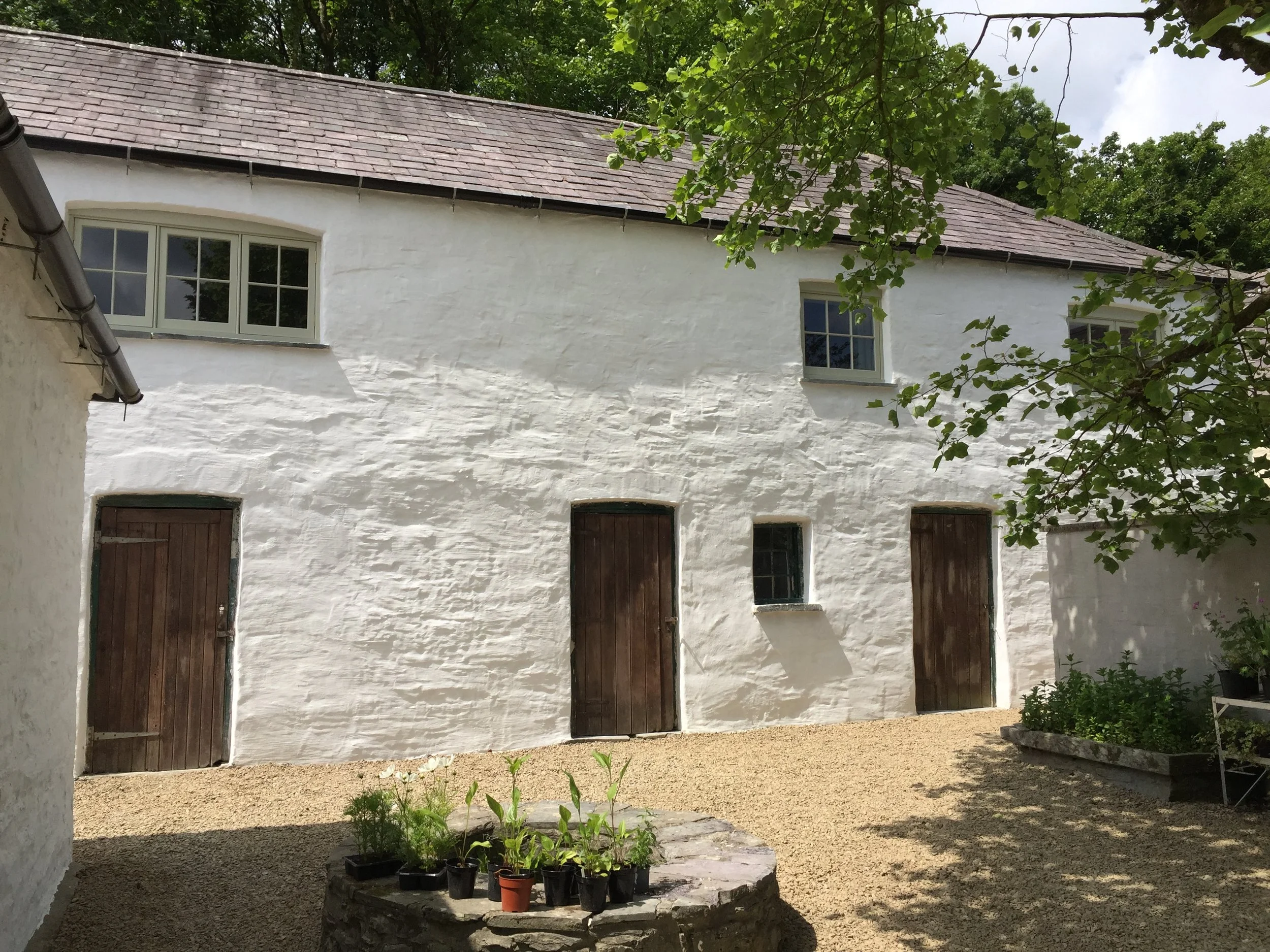 White cottage with three wooden doors and two windows, surrounded by trees and a gravel yard, with potted plants on a stone ledge in the foreground.