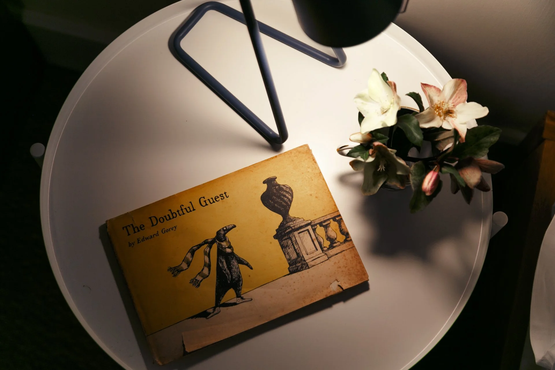 A white round table with a vintage book titled 'The Doubful Guest' by Edward Gorey, and a small vase with white and pink flowers. Part of a lamp with a blue stand is also visible.