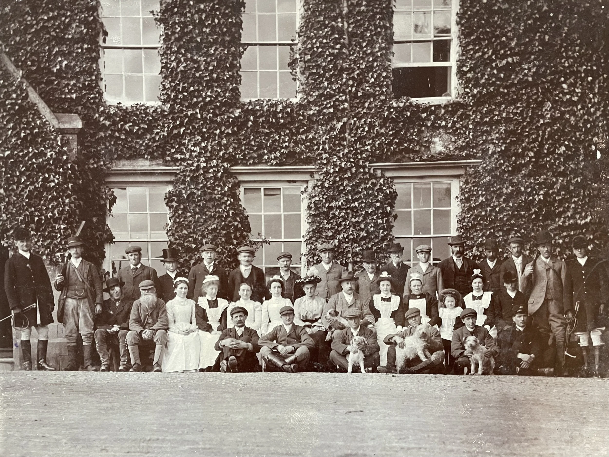 A large group of men, women, and children posing outdoors in front of a vine-covered building in a black-and-white photograph.