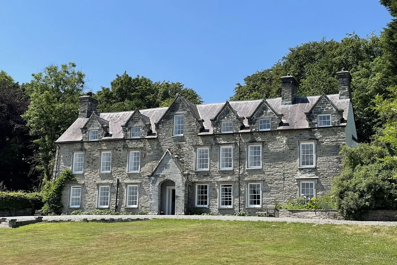Large stone house with multiple windows, dormer windows on the roof, and surrounded by green trees and grass.