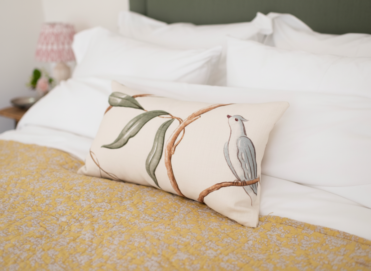 Decorative pillow with a bird and branch design on a bed with white sheets and a yellow blanket in a bedroom.
