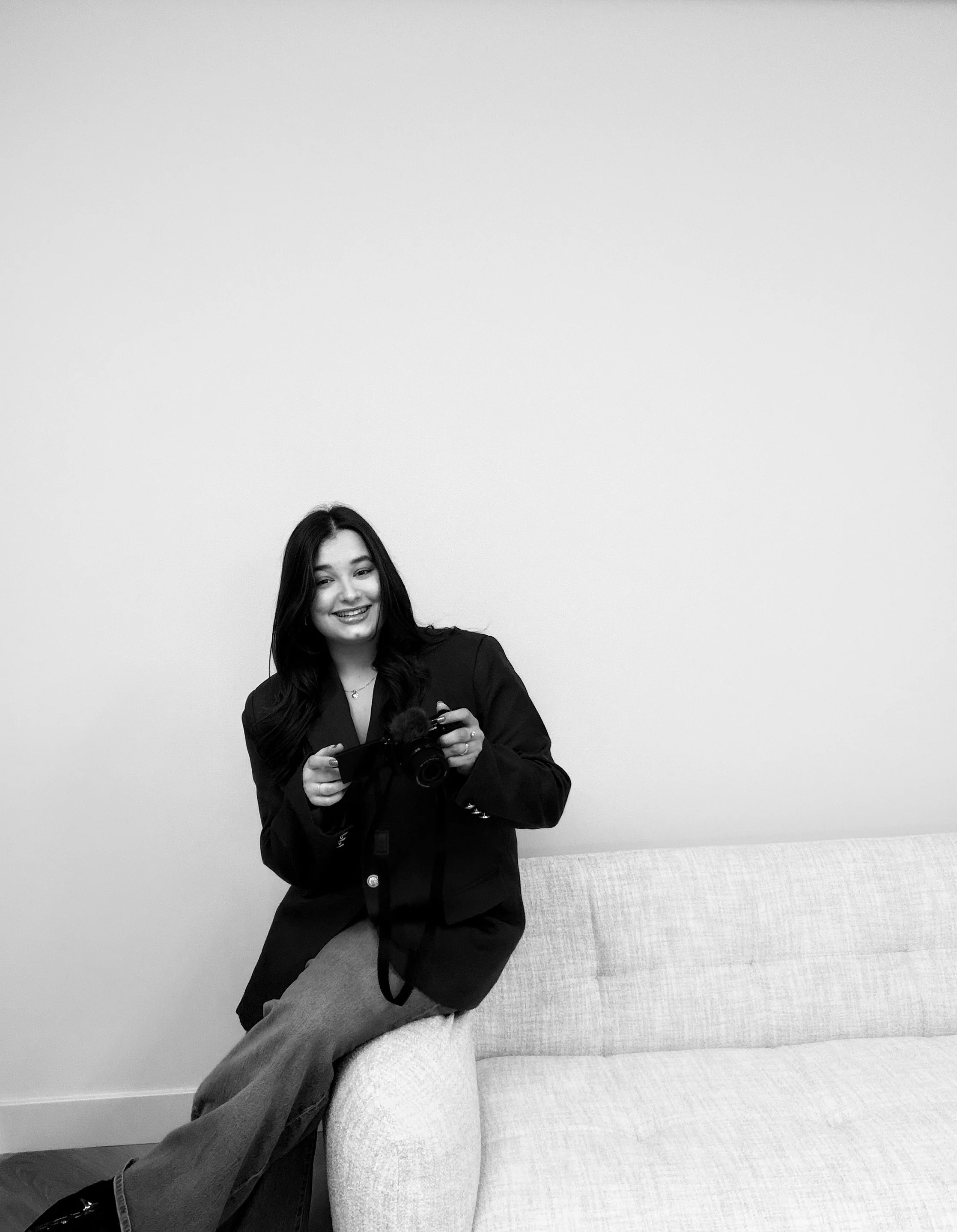 Een lachende vrouw met lang haar zit op de armleuning van een lichtgekleurde bank, terwijl ze een camera vasthoudt. De achtergrond is een effen witte muur.