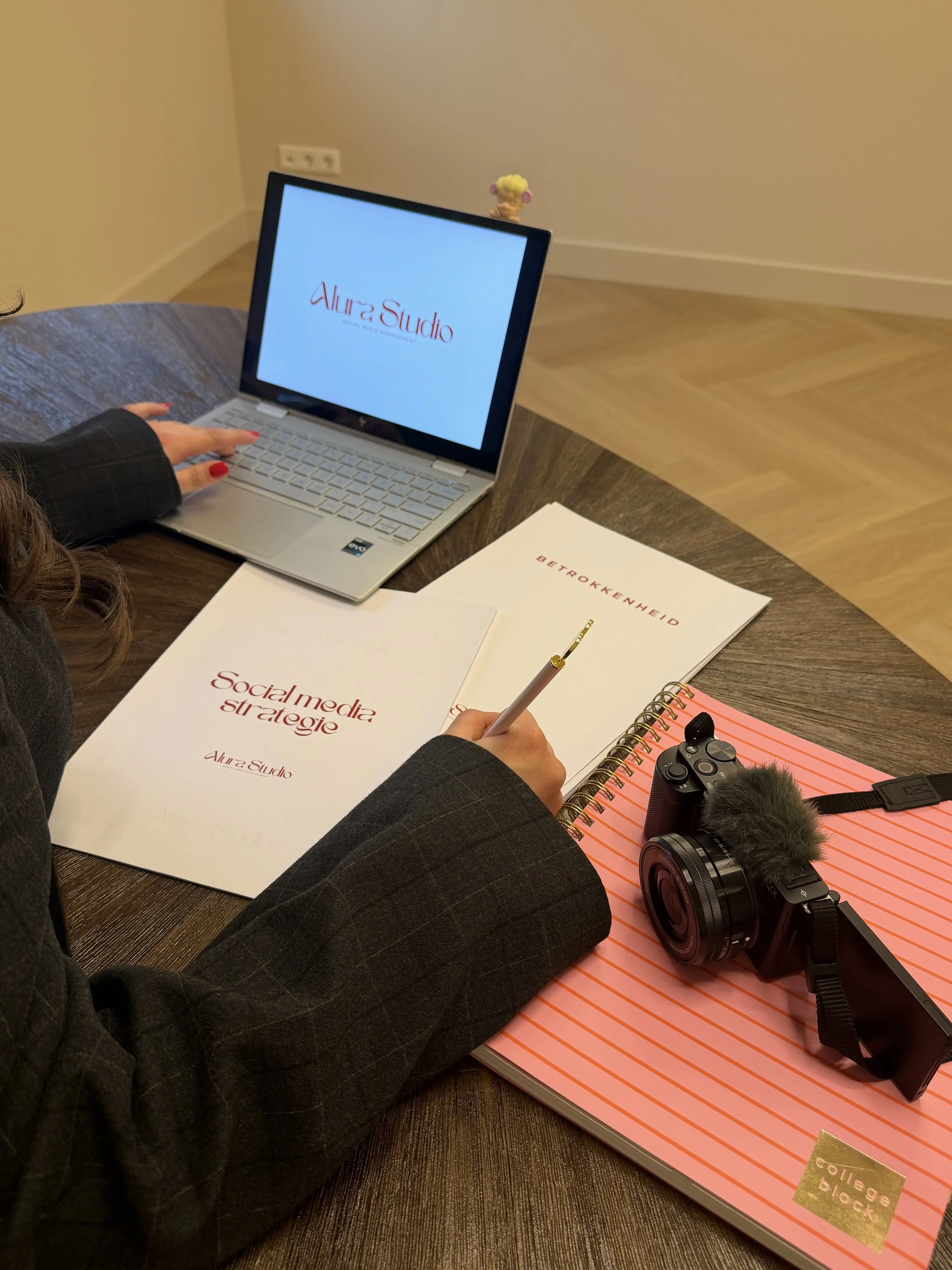 Een persoon werkt aan een bureau met een laptop, notitieboekjes, een camera met microfoon, en een paar documenten met teksten, terwijl ze op een stoel zit.