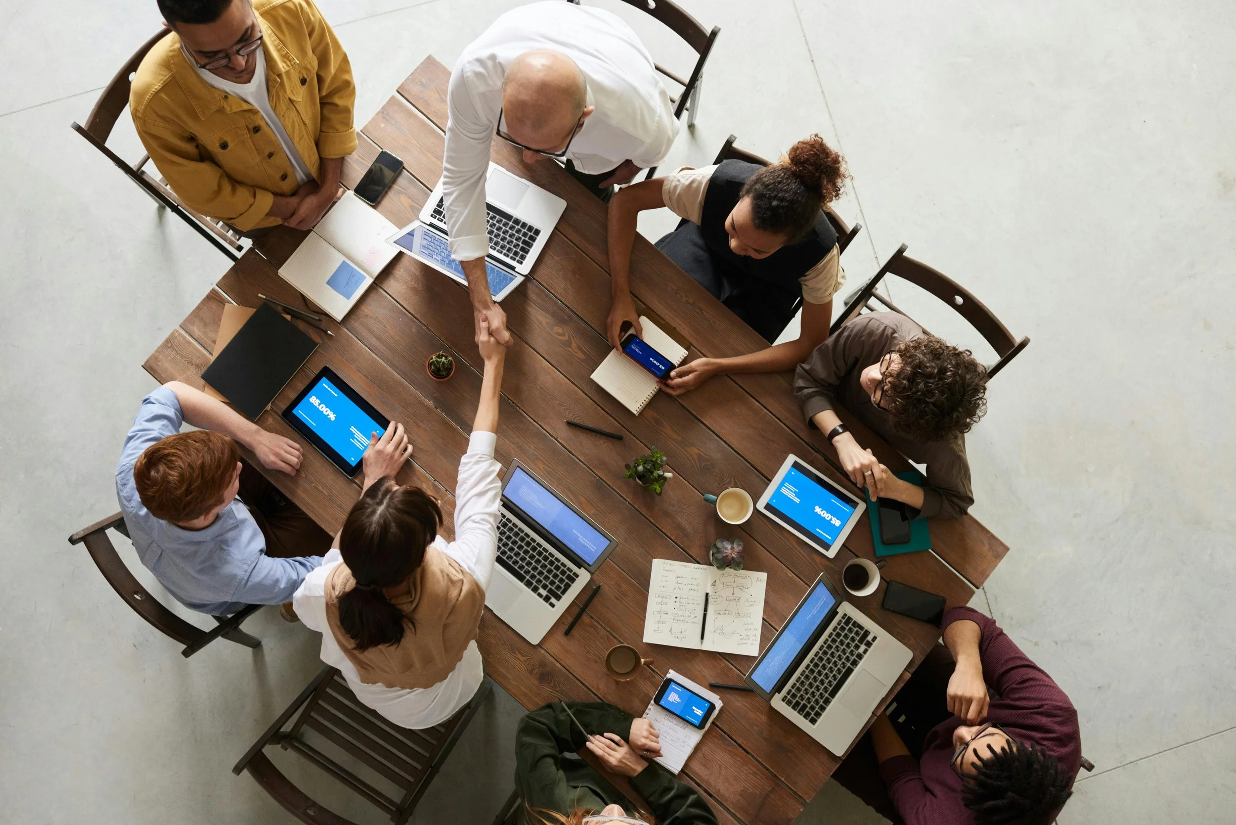 A group of eight diverse people around a wooden conference table working on laptops, tablets, and notebooks. Some are shaking hands, papers and coffee cups are on the table, and the setting is a modern, open workspace.