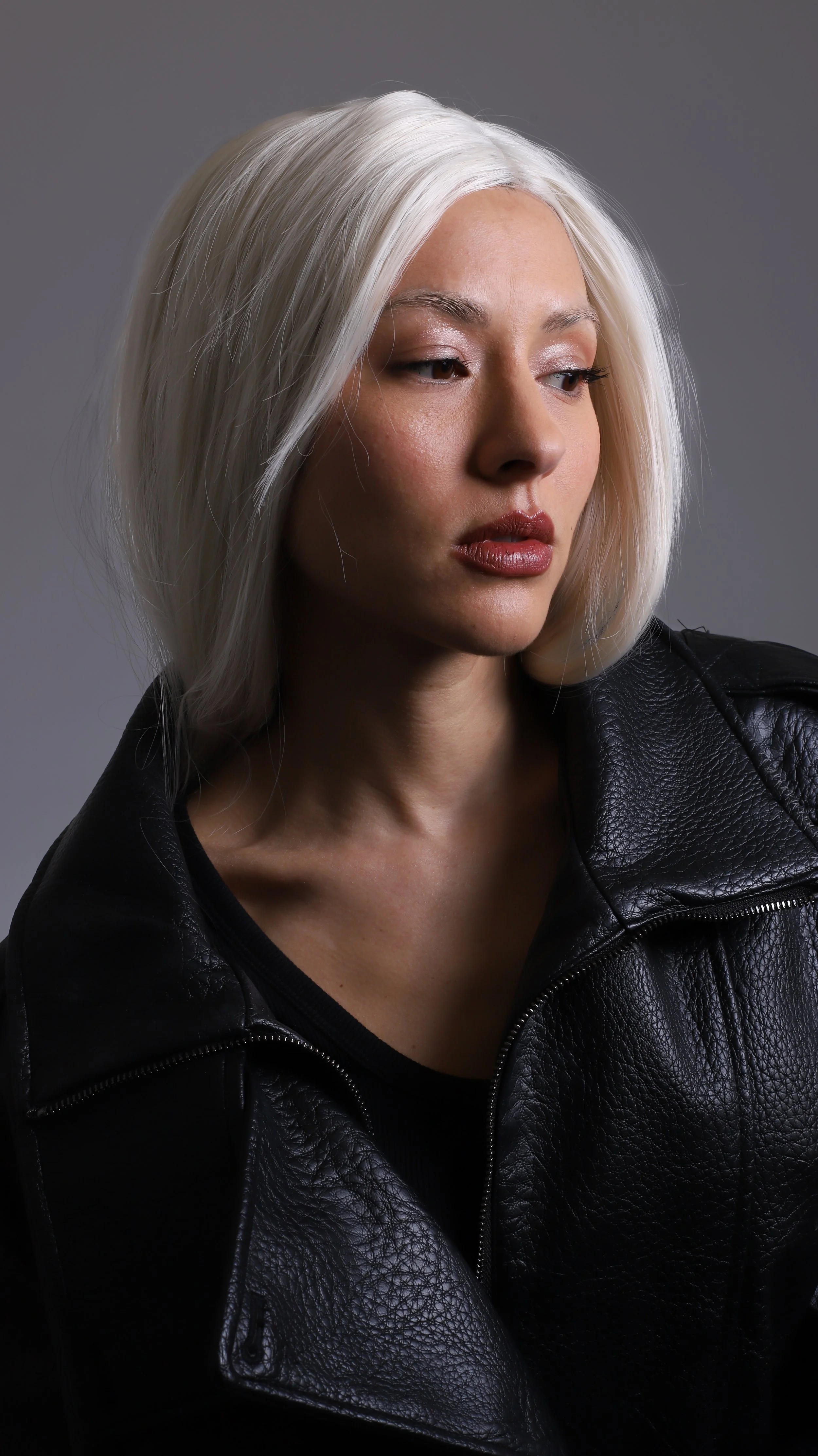 A woman with platinum blonde hair wearing a black leather jacket, looking slightly to the side against a gray background.