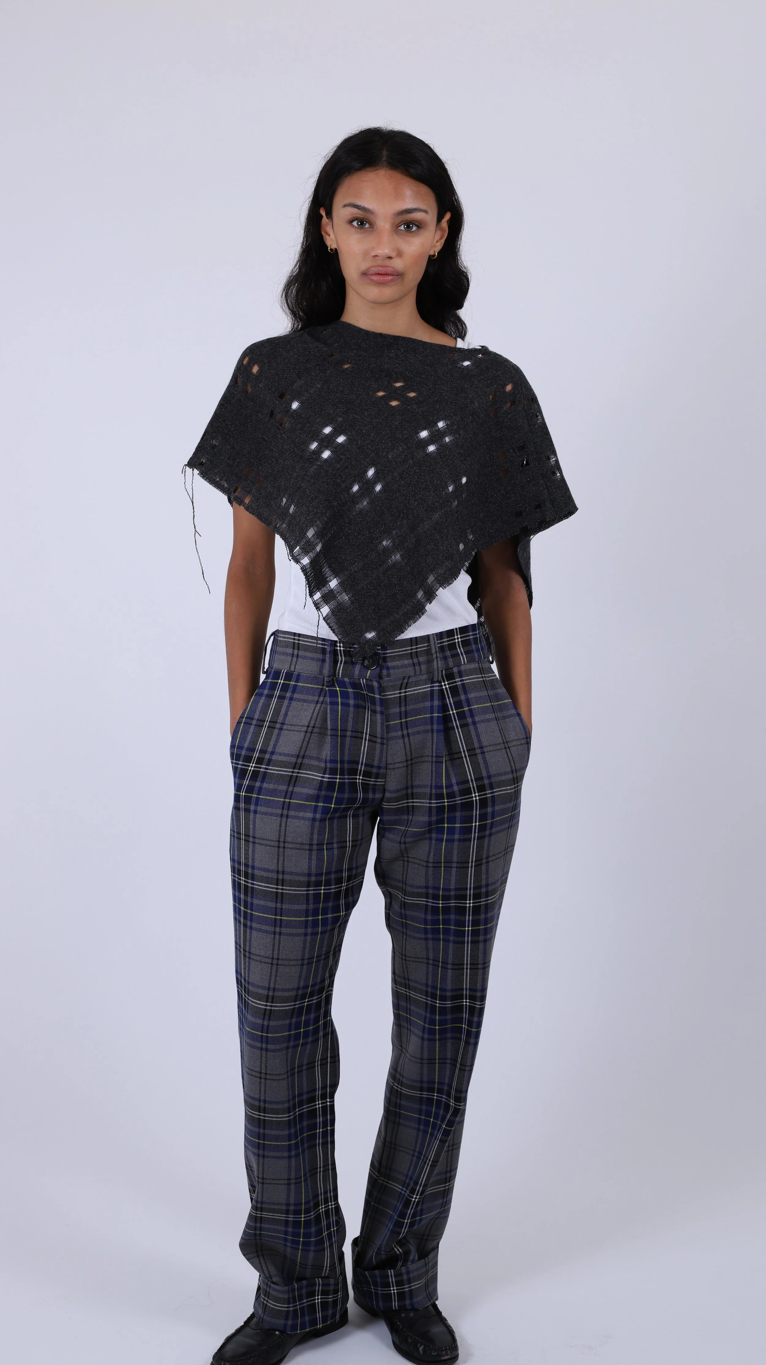 A woman with dark hair, wearing a black checkered shawl over a white top and plaid pants, standing against a plain white background.