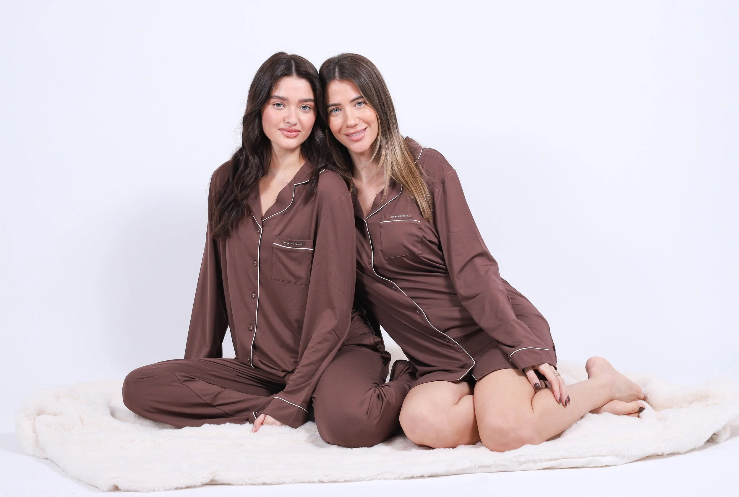 Two women sitting on a white furry blanket, wearing matching brown satin pajamas, smiling at the camera against a plain white background.