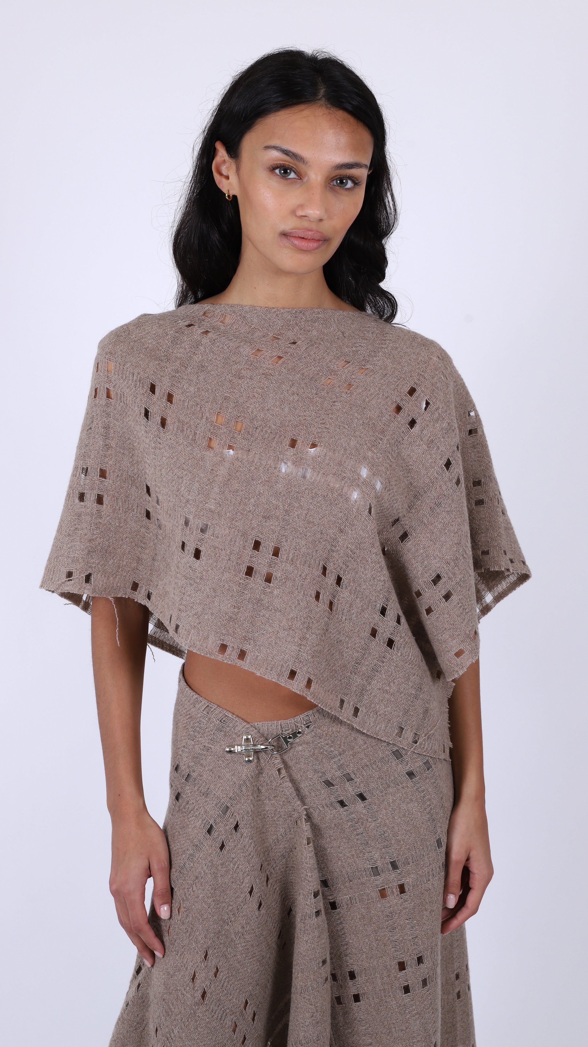 A woman with dark wavy hair and earrings wearing a matching beige knitted top and wide-leg pants with cut-out patterns, standing against a plain white background.