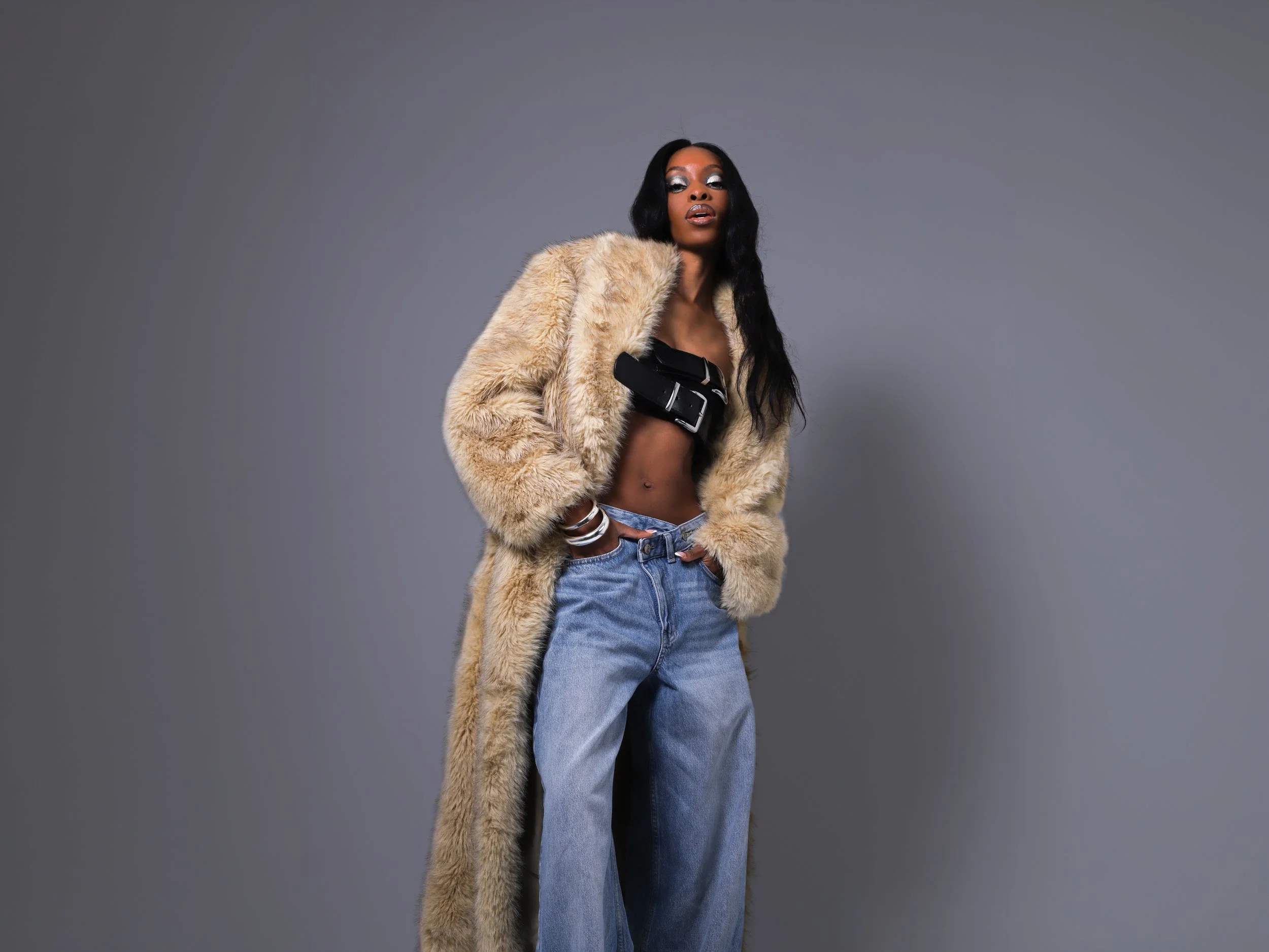 A woman with long black hair, wearing a beige fur coat, black crop top, and loose blue jeans, standing against a gray background.