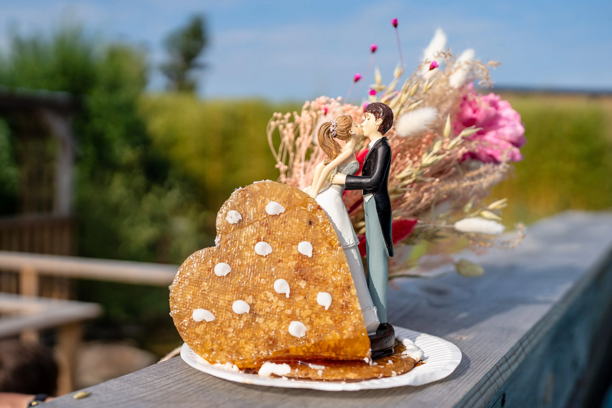 Une pièce montée avec des figurines de mariés et un gâteau en forme de cœur, décorée de fleurs séchées, placée sur une table en bois extérieure.
