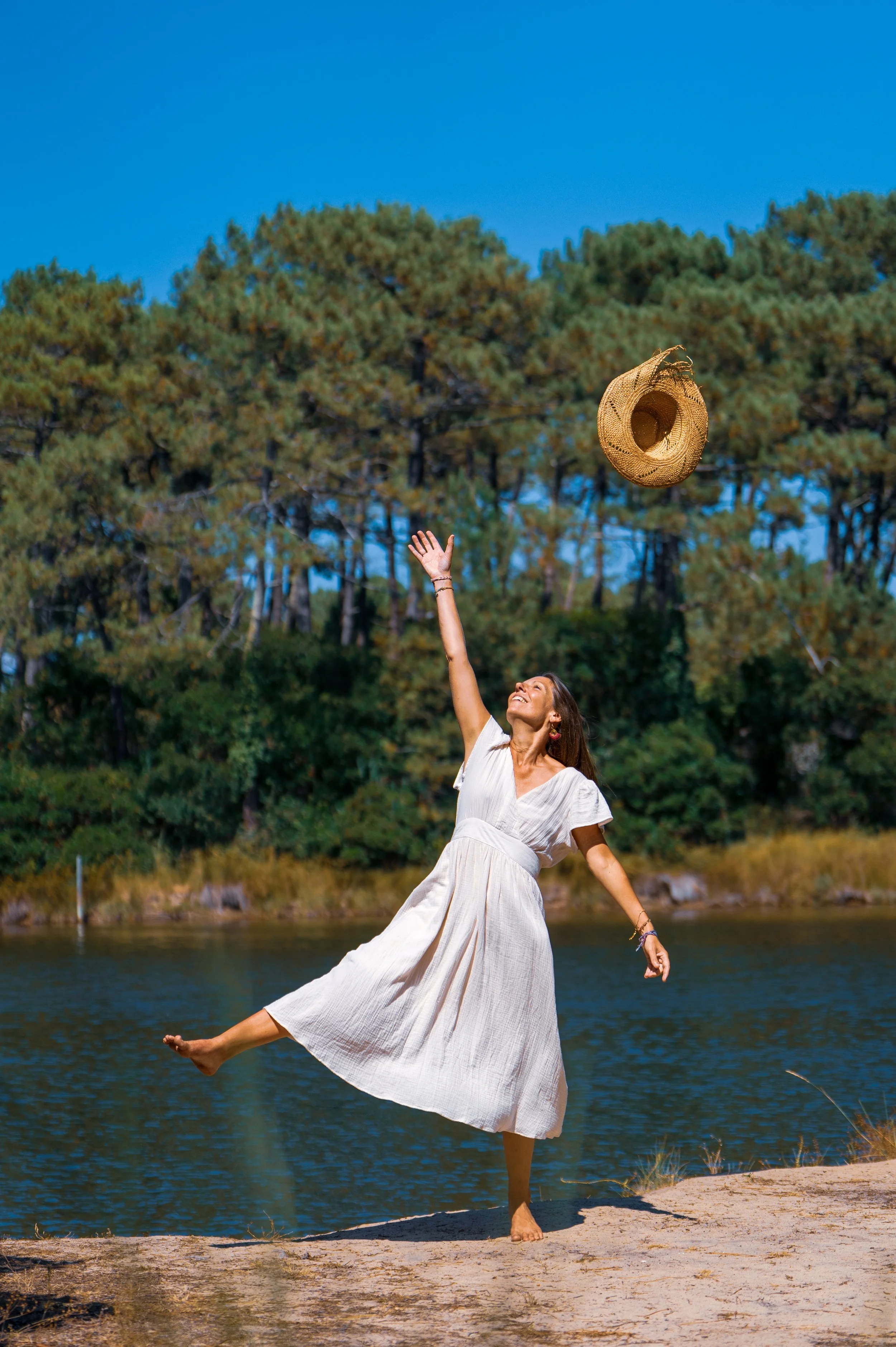 Femme en robe blanche lançant un chapeau en paille en l'air près d'un lac avec des arbres en arrière-plan.